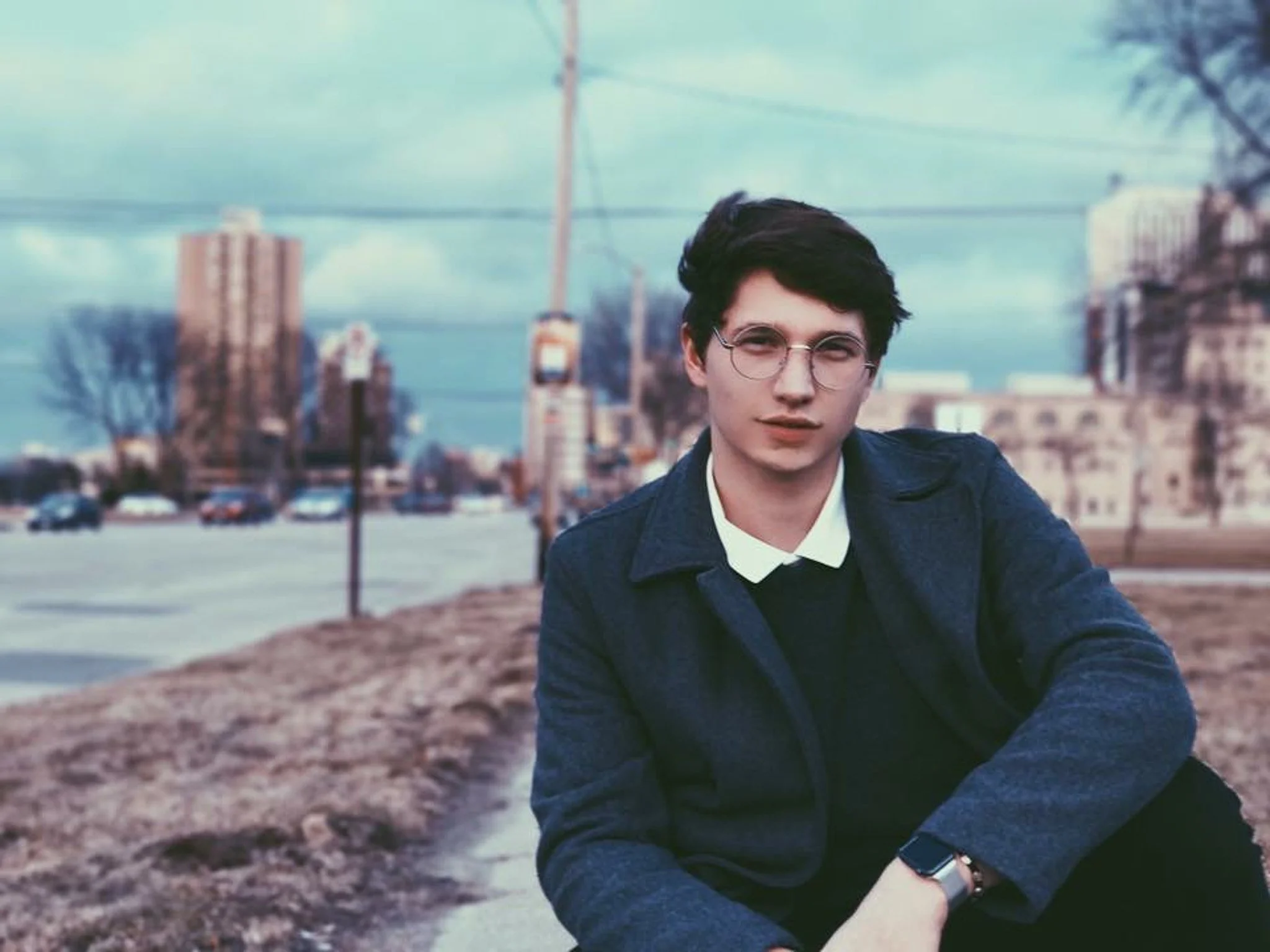 Young man with dark hair and glasses wearing a black jacket and white shirt, sitting outdoors with a cityscape and water in the background.