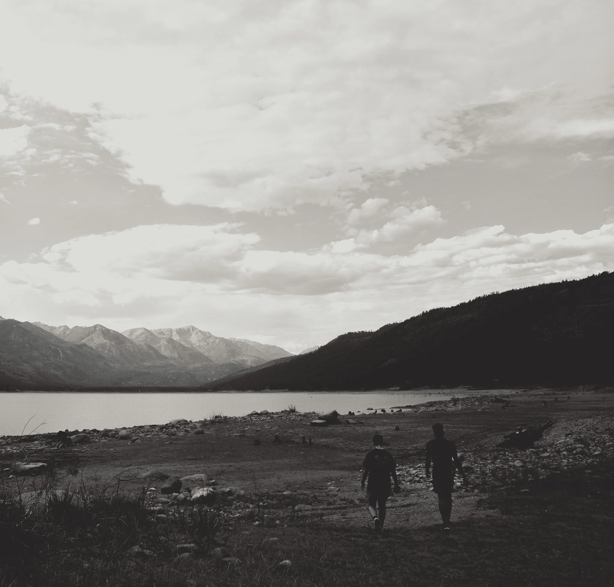 Black and white photo of a river with mountains in the background and people walking by the riverbank.