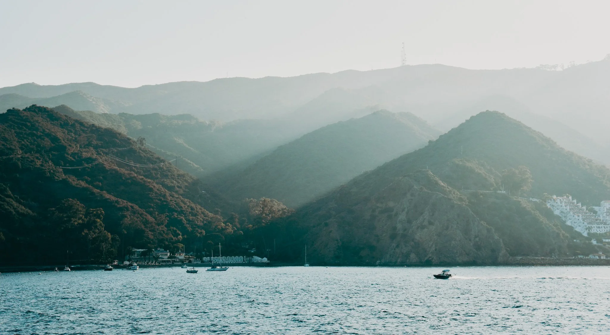 Hilly coastal landscape with lush green mountains, a few boats on the water, and a small town at the shoreline under a hazy sky.