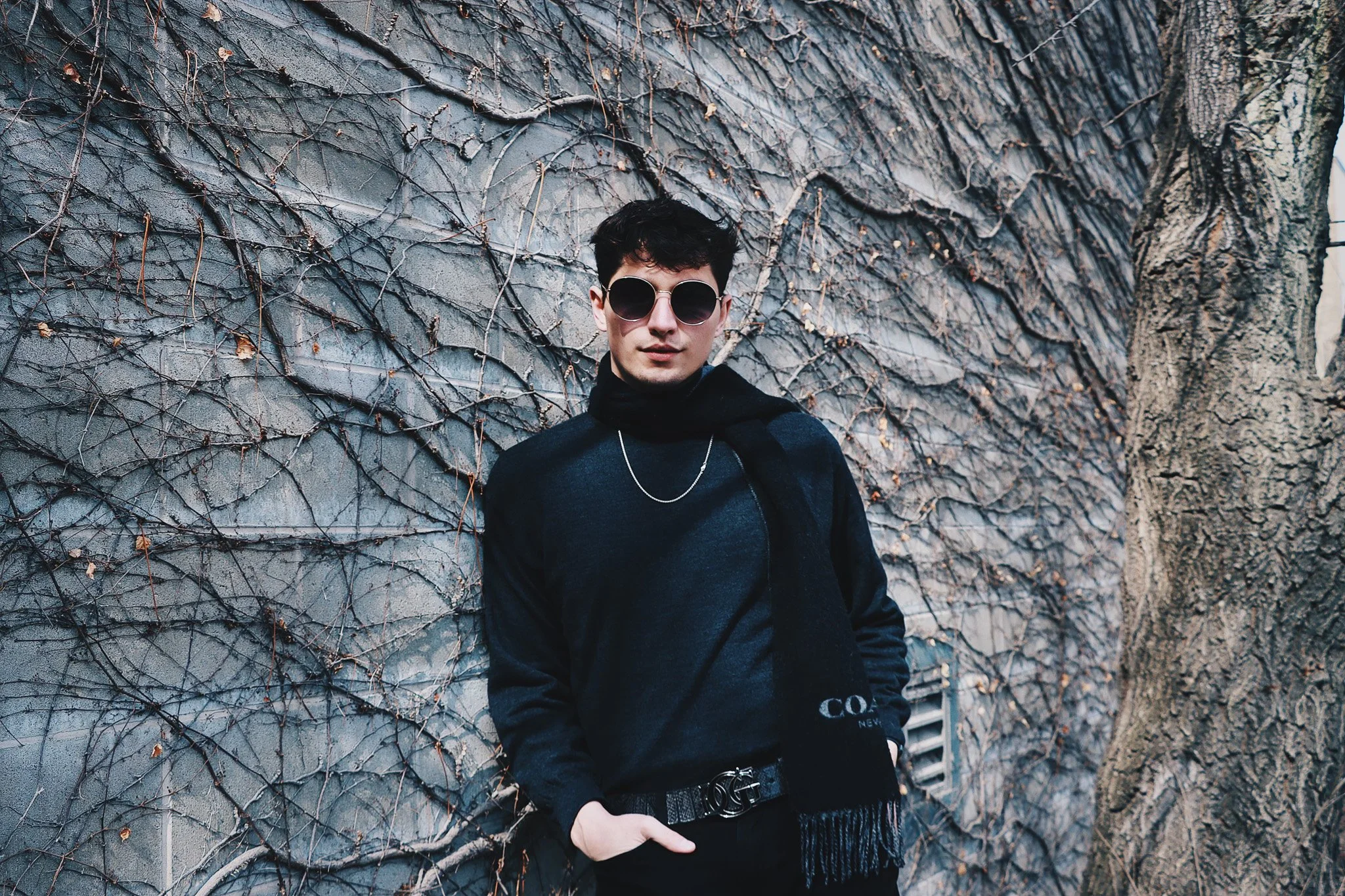 A young man with dark hair and sunglasses standing in front of a wall covered in vines and textured concrete.