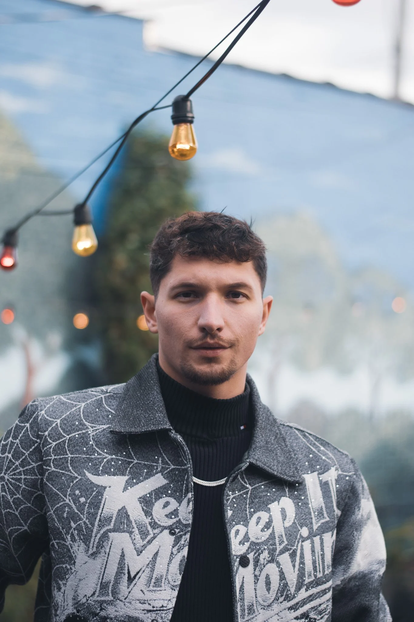 Young man with short dark hair and slight facial hair wearing a black turtleneck and a gray patterned jacket with white text, standing outdoors under decorative string lights with blurred trees and sky in the background.