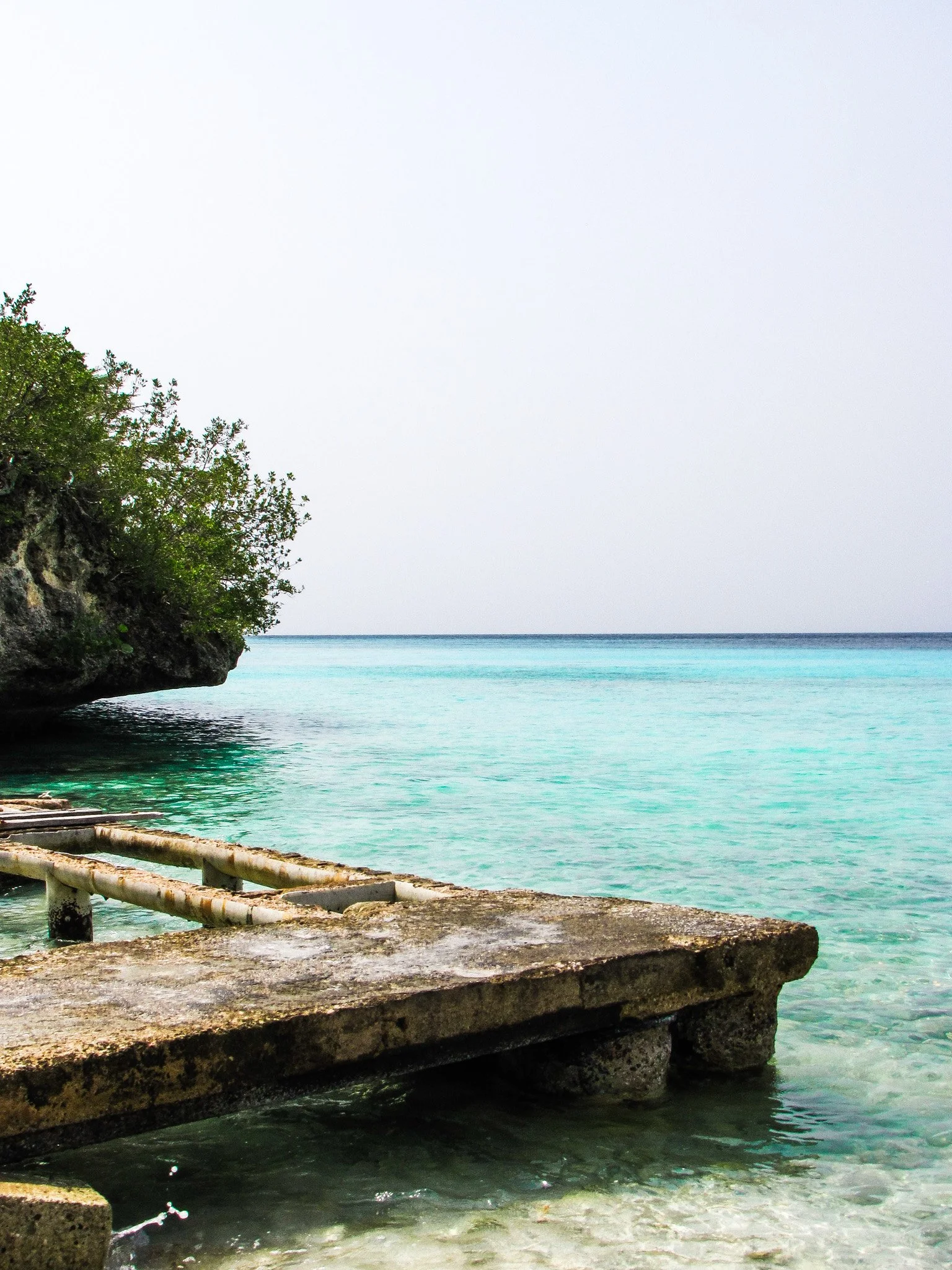 A rocky shoreline with a concrete slab, green shrubbery on a cliff, and calm turquoise ocean waters extending to the horizon under a pale sky.