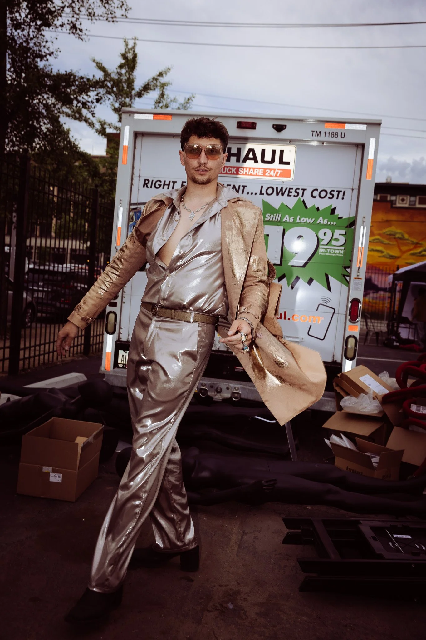 A person dressed in shiny, metallic silver clothing, including a blazer and wide-leg pants, standing in front of a U-Haul truck with promotional signage. The person is wearing sunglasses, a necklace, and has short, curly hair. There are open boxes an