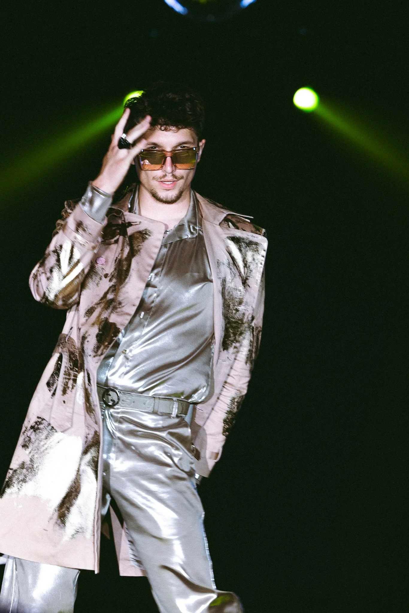 A man with curly dark hair and sunglasses wearing a shiny silver shirt, patterned beige coat, and silver pants, standing against a black background with green stage lights.