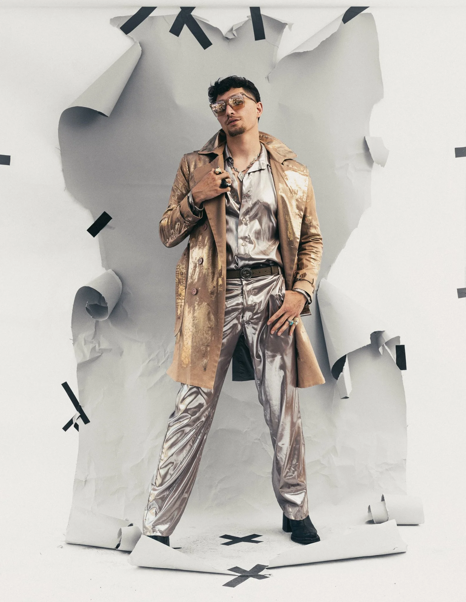 Stylish man wearing sunglasses, metallic jacket, and shiny pants standing against torn white paper background with black tape