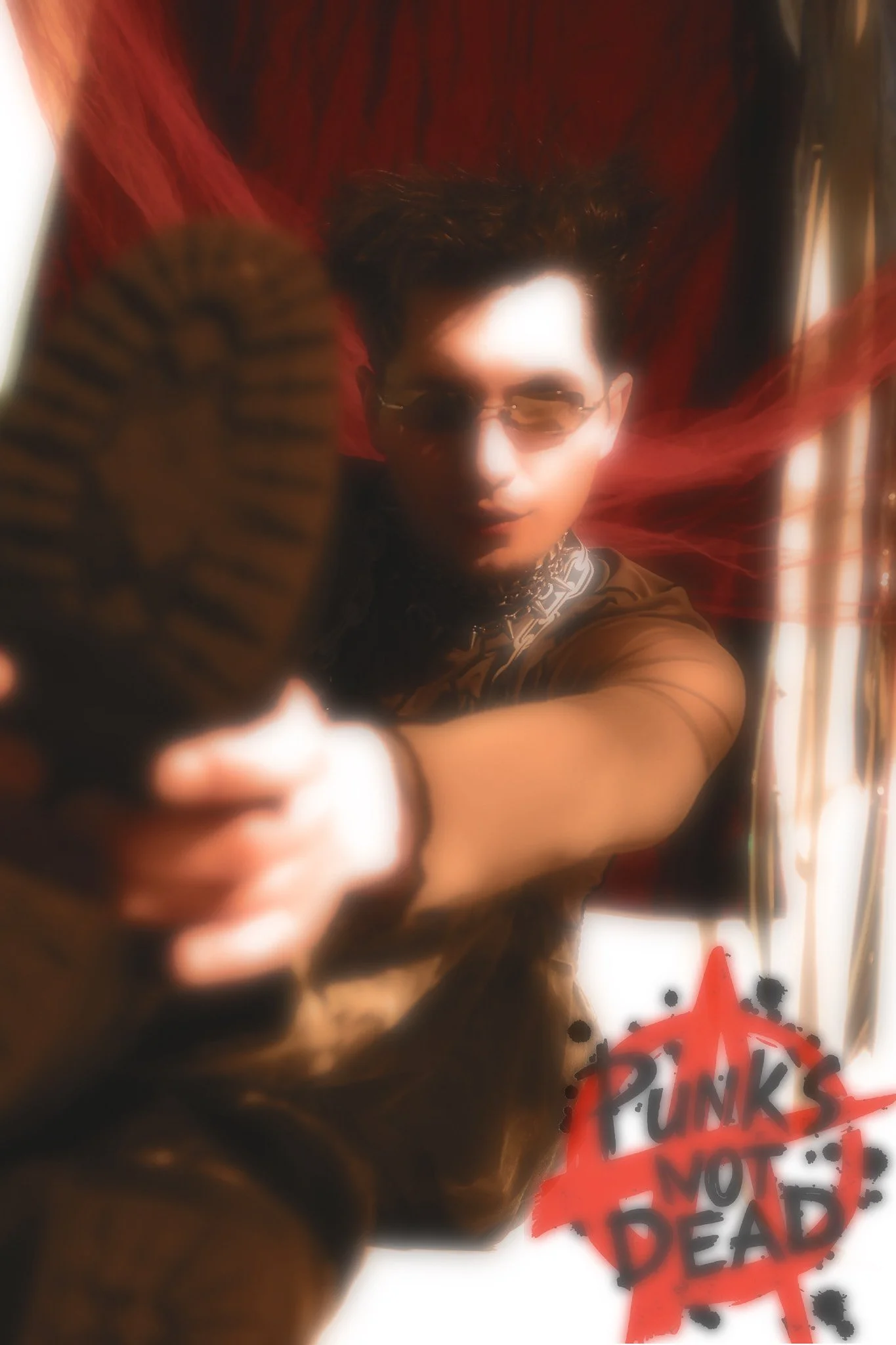 A person with dark hair and sunglasses holds a spray paint can, spray painting a red anarchy symbol and words such as 'F***' and 'Dead' on a white surface, with red fabric in the background.