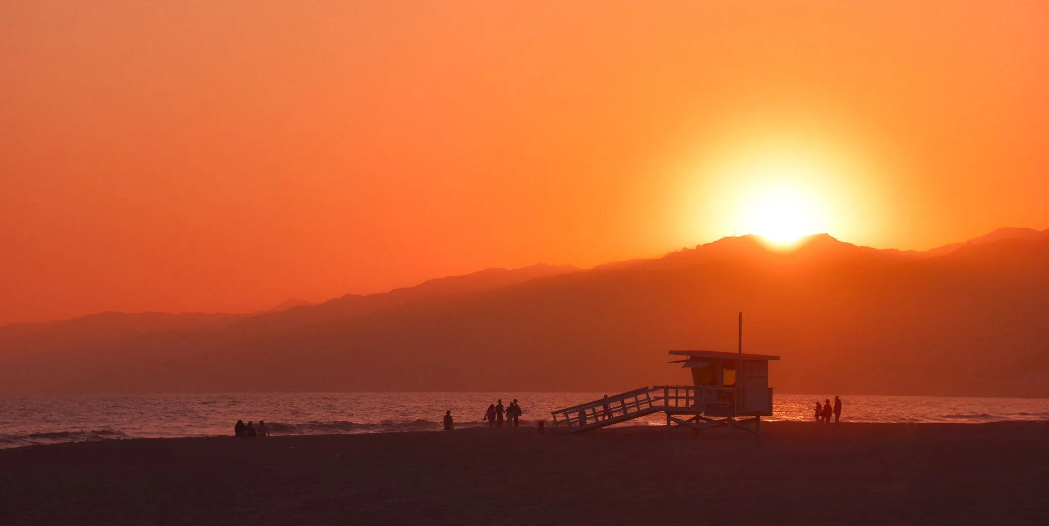 Sunset over the ocean with silhouettes of people on the beach, a lifeguard tower, and mountains in the background.