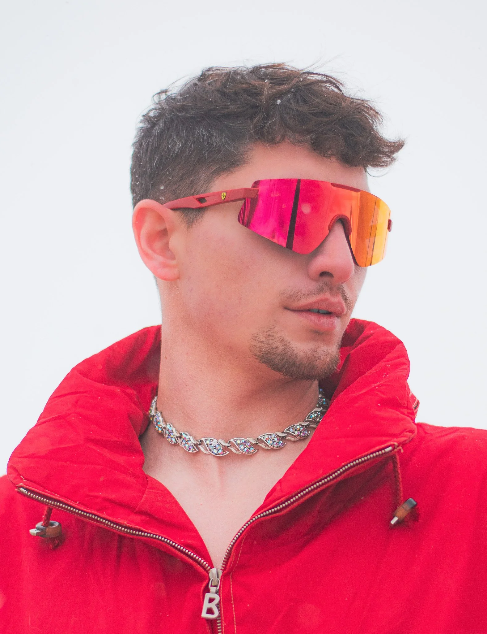 A man wearing a bright red jacket and reflective sports sunglasses with orange mirror lenses, standing outdoors in snowy weather, with snowflakes on his hat.