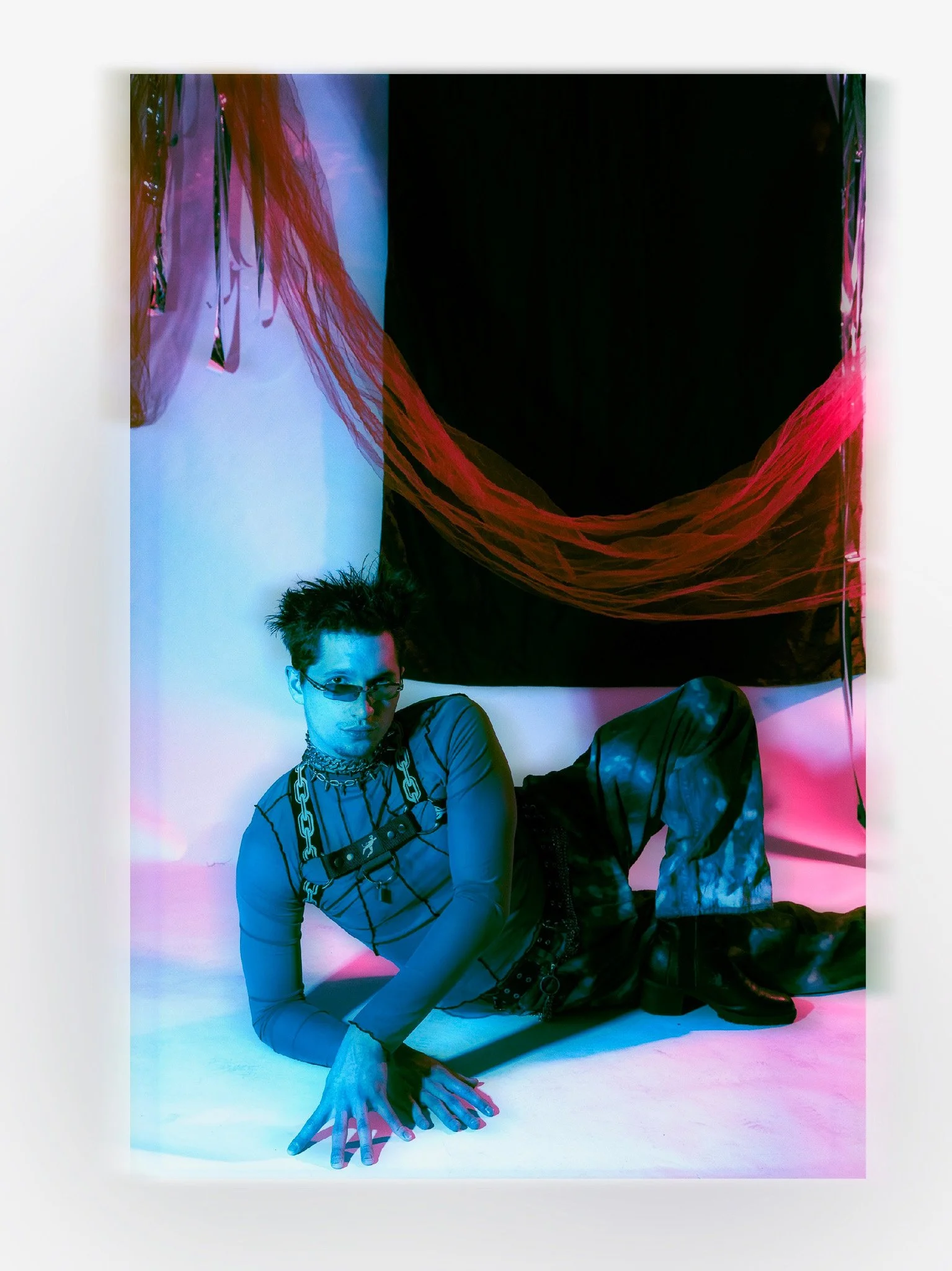 A young person with short dark hair and glasses, wearing a striped shirt with chain accessories, is posed lying on the floor with one hand touching the ground and the other hand gripping the floor, in front of a large, dark-colored abstract painting 