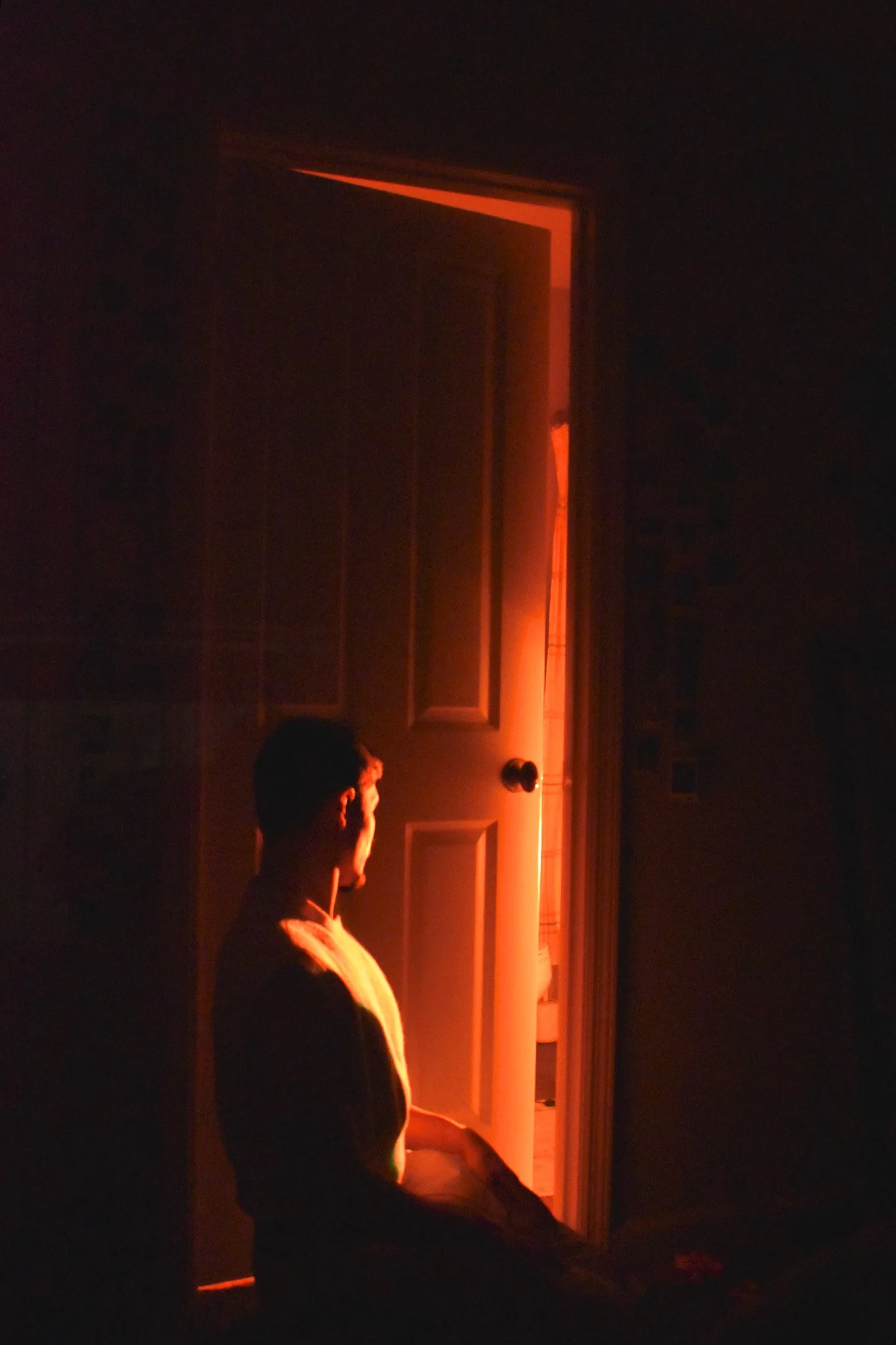 A dark room with a person standing near a door illuminated by warm light. The person is facing the door, and their profile is visible, with short dark hair. The door is slightly open, revealing a hint of the lit space beyond.