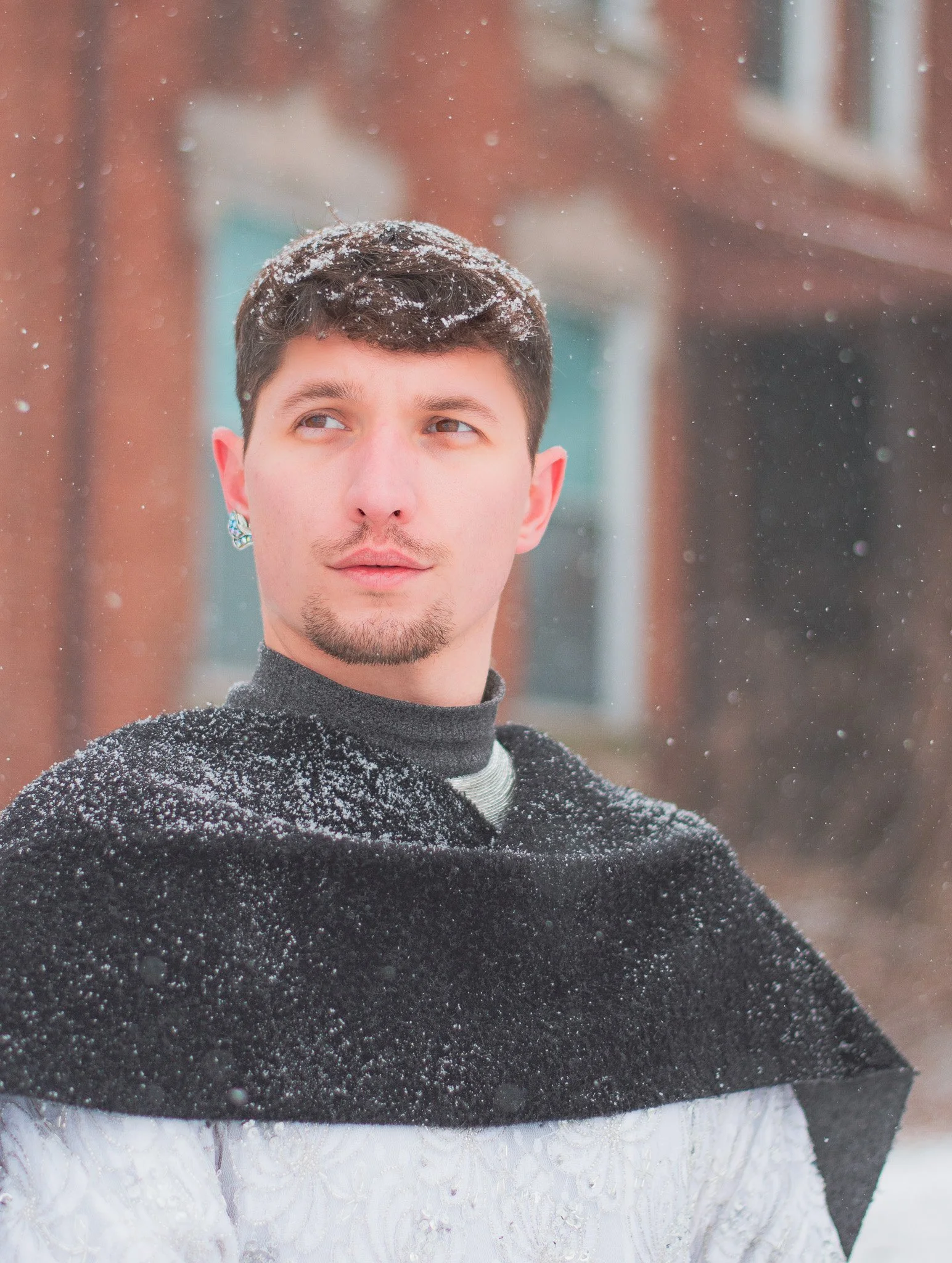 Close-up of a young man with short dark hair, facial hair, and earrings, in a snowy outdoor setting, wearing a dark sweater with snow on his shoulders, gazing to the side with a thoughtful expression.