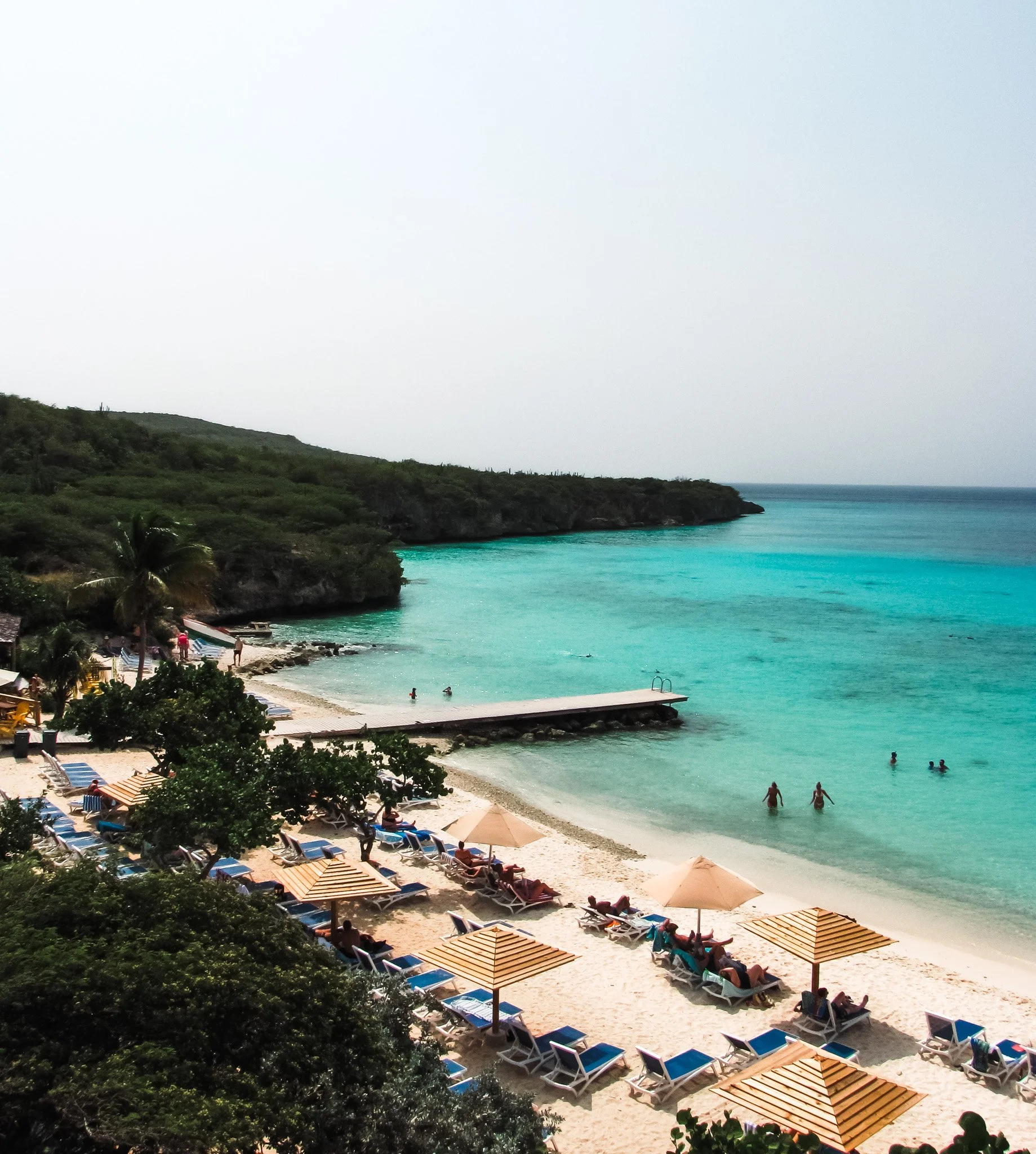 Beach scene with turquoise water, sandy shore, lounge chairs, umbrellas, and people swimming and relaxing. Green hills and trees frame the shoreline under a cloudy sky.