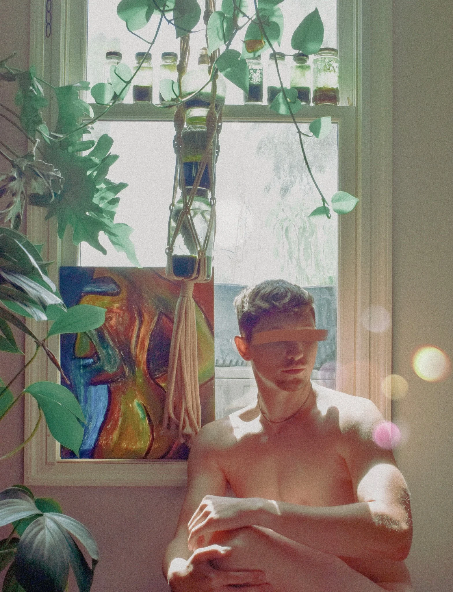 A person with short hair taking a mirror selfie, with mountains of hair on their arm, in a room with a window. There are plants and a colorful painting in the background, and bottles with plants on the window sill.