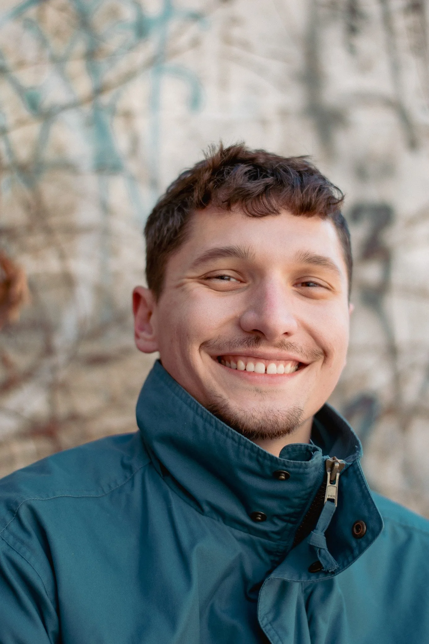 Close-up of a young man smiling, wearing a dark blue jacket, standing outdoors with a blurred stone wall background.