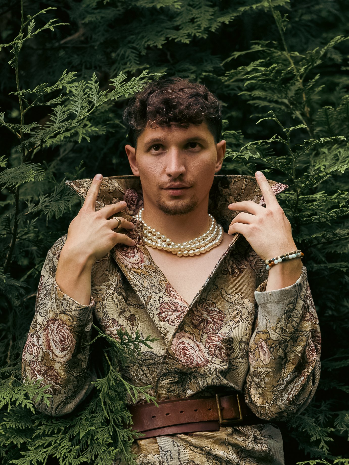 A person with short, curly hair and a goatee, posing among greenery. They are wearing a patterned blazer, pearl necklaces, and bracelets, and are holding the collar of their blazer with both hands.