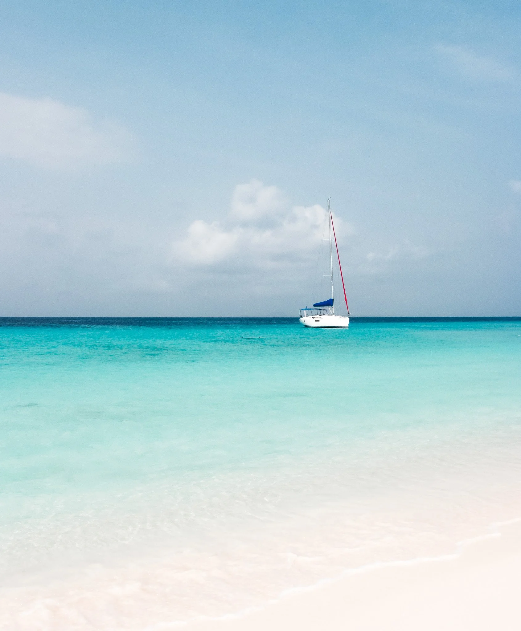A sailboat with a red mast and blue canopy floating on clear, turquoise ocean water under a partly cloudy sky.