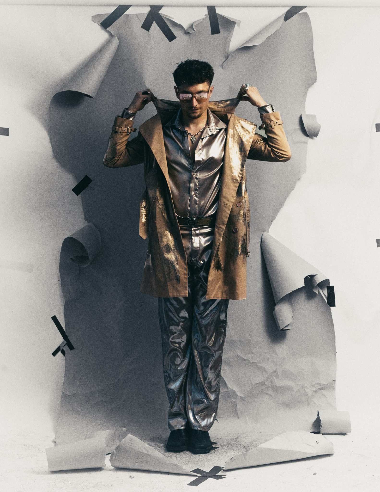 A man with dark curly hair wearing reflective glasses, metallic clothing, and a layered trench coat, stands with hands on shoulders against a torn paper backdrop with black tape. The torn paper creates a textured frame around him.