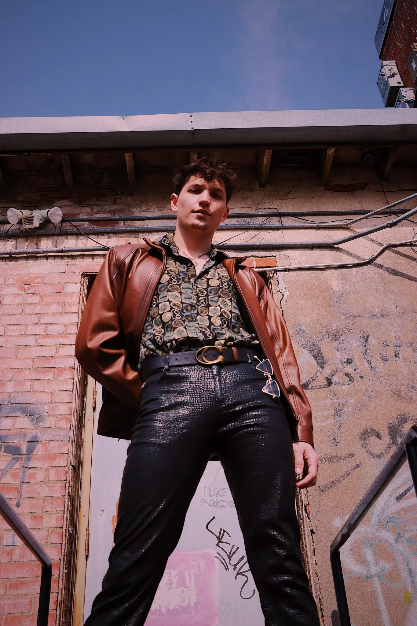 A young man with dark hair standing outdoors in front of a brick wall and a graffiti-covered door, wearing a patterned shirt, black shiny pants, a brown leather jacket, and sunglasses hanging from his pants belt. He looks confident and relaxed, with 