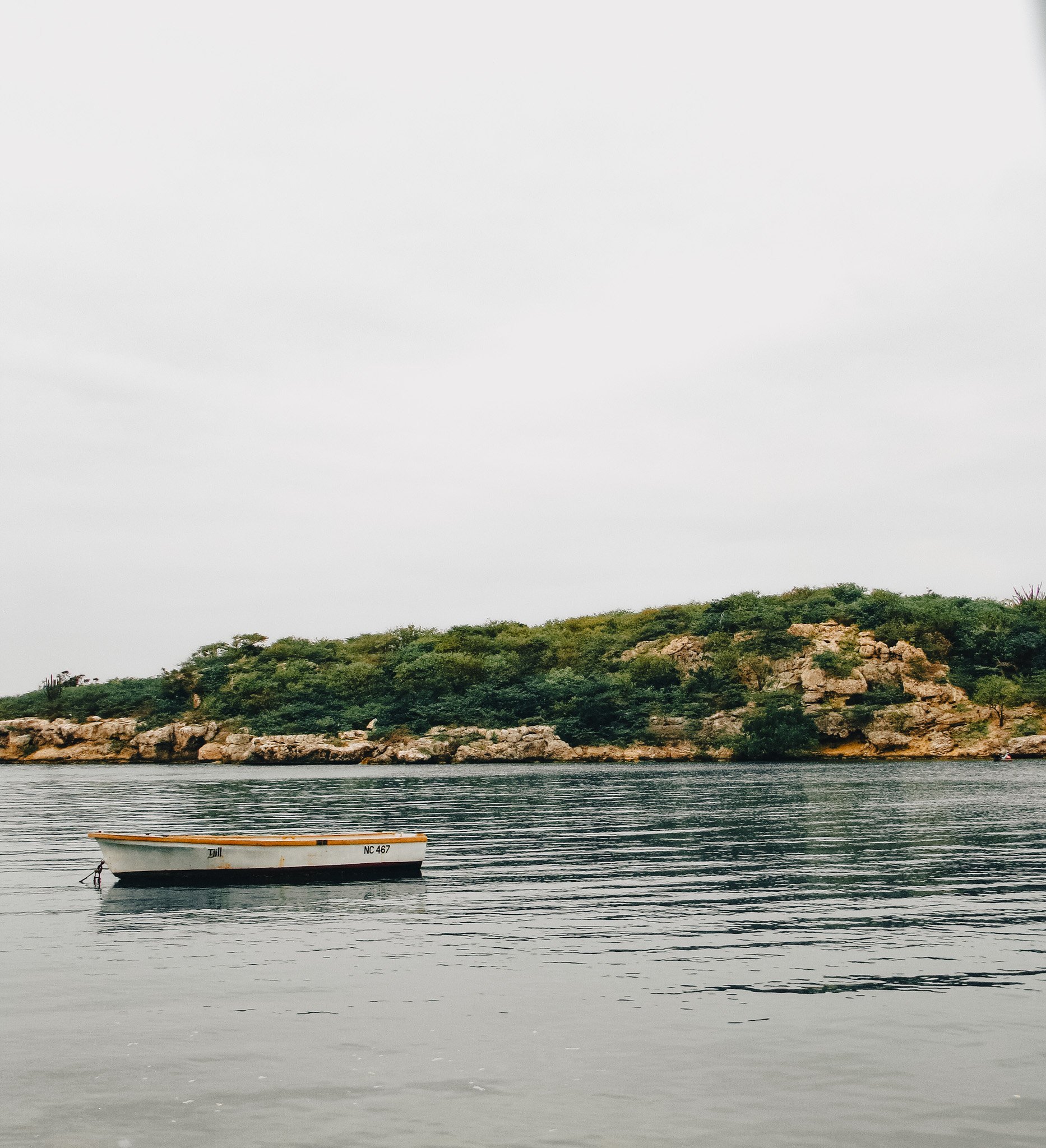 A white boat floating on calm water with a rocky and green hillside in the background under a cloudy sky.