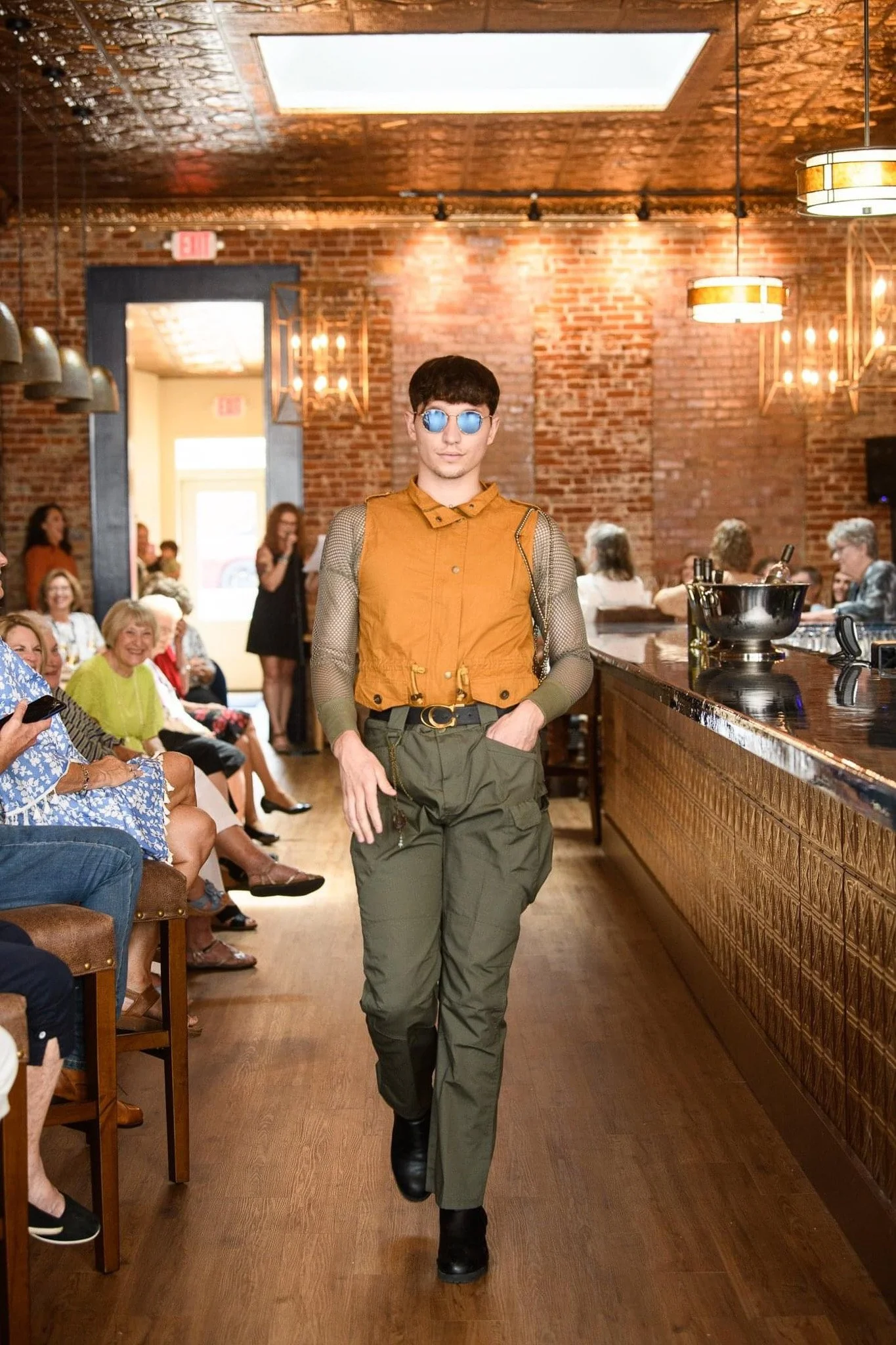 A person wearing sunglasses, an orange vest, mesh sleeves, and green pants walking through a crowded bar or restaurant with brick walls and warm lighting.