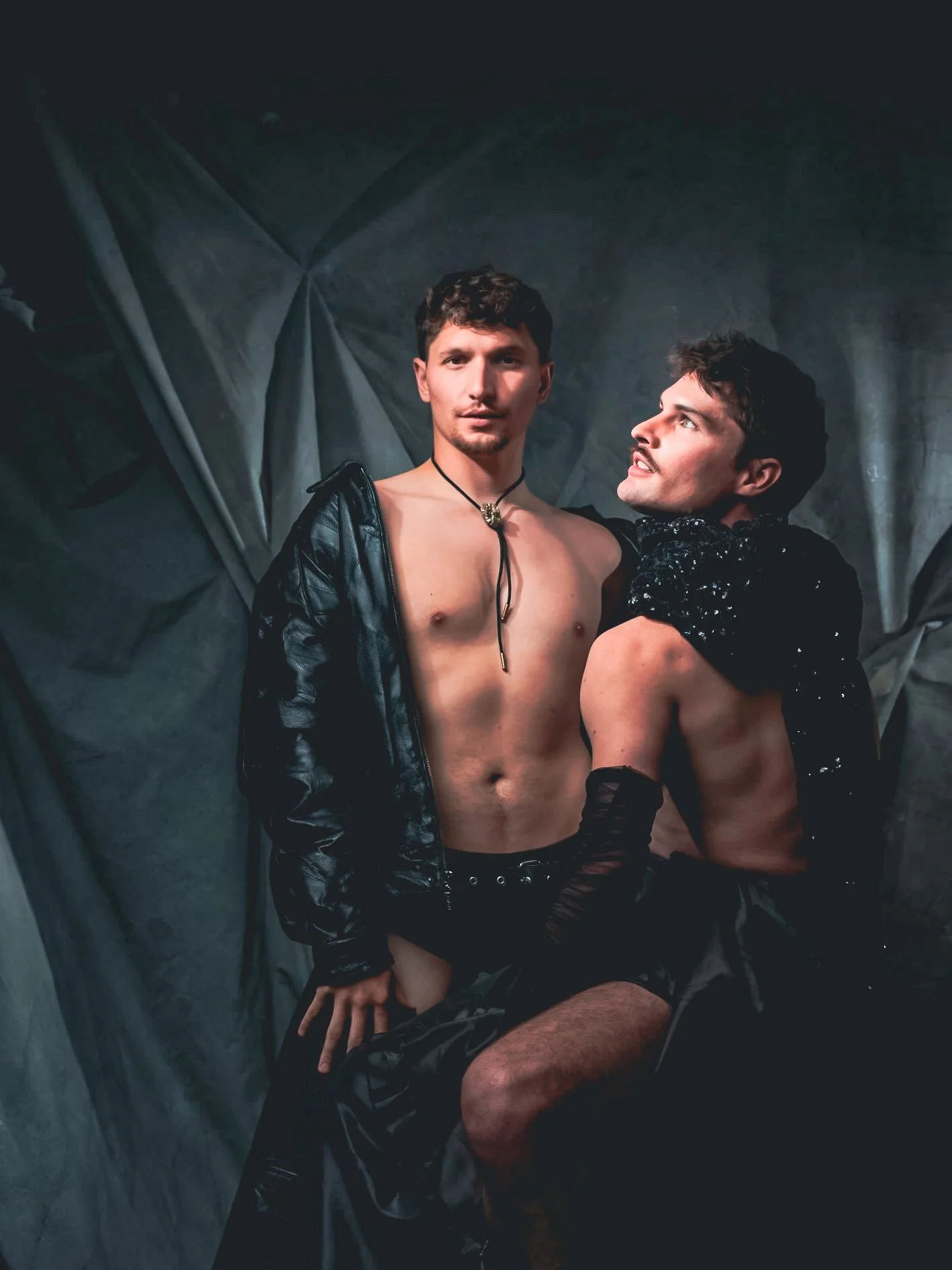 Two shirtless men with dark, curly hair and athletic builds posing with serious expressions. One wears black gloves and a leather jacket slightly off his shoulders, the other looks at the first man, wearing black sequin fabric on his arms and waist. 