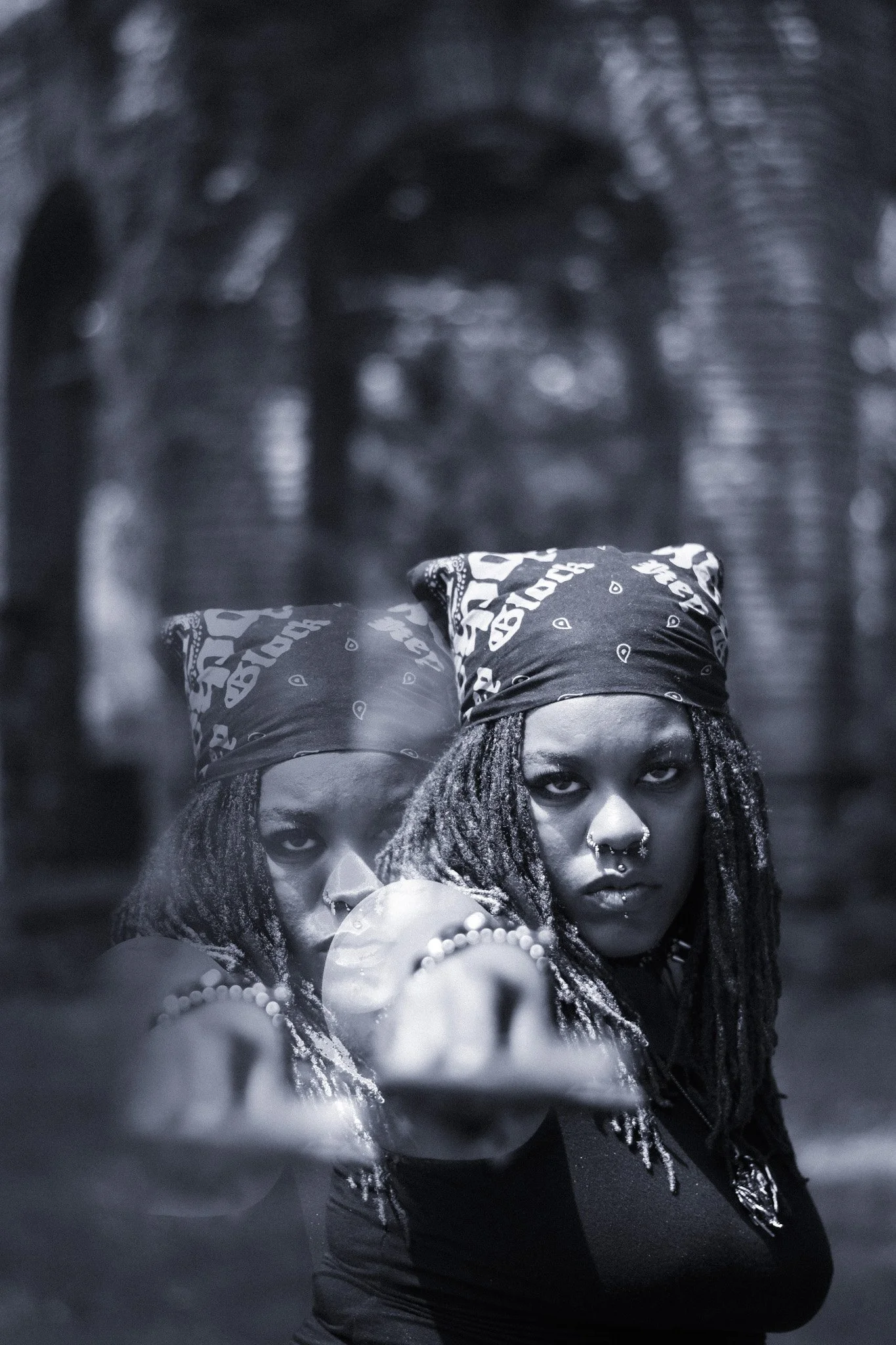 A person with long dreadlocks, nose and septum piercings, and a bandana on their head, extending their arm towards the camera with a serious expression, dark background and blurred figures.