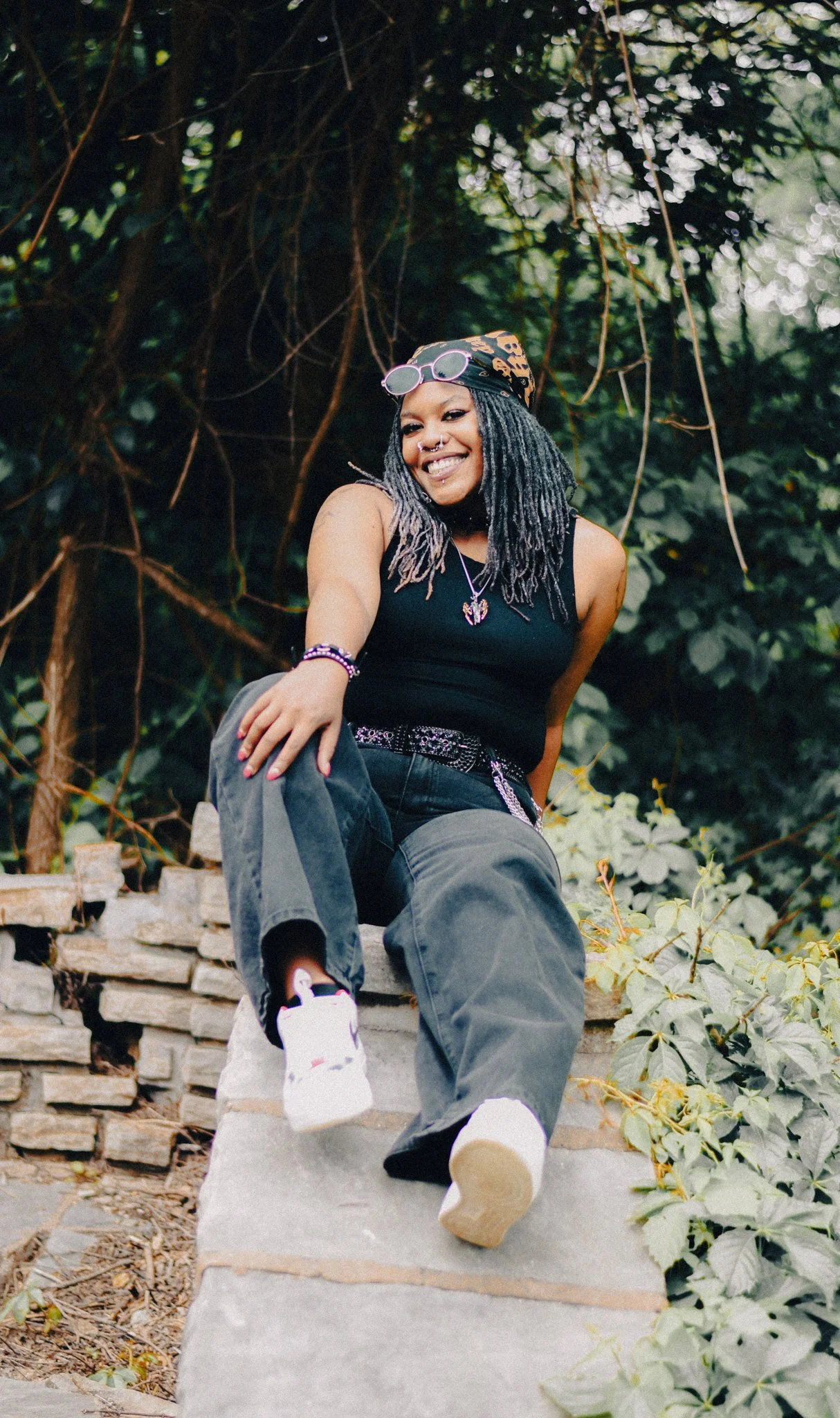A woman with dreadlocks, sunglasses on her head, and a bandana, sitting on a stone step outdoors, smiling, wearing a black tank top, jeans, and sneakers, surrounded by greenery.