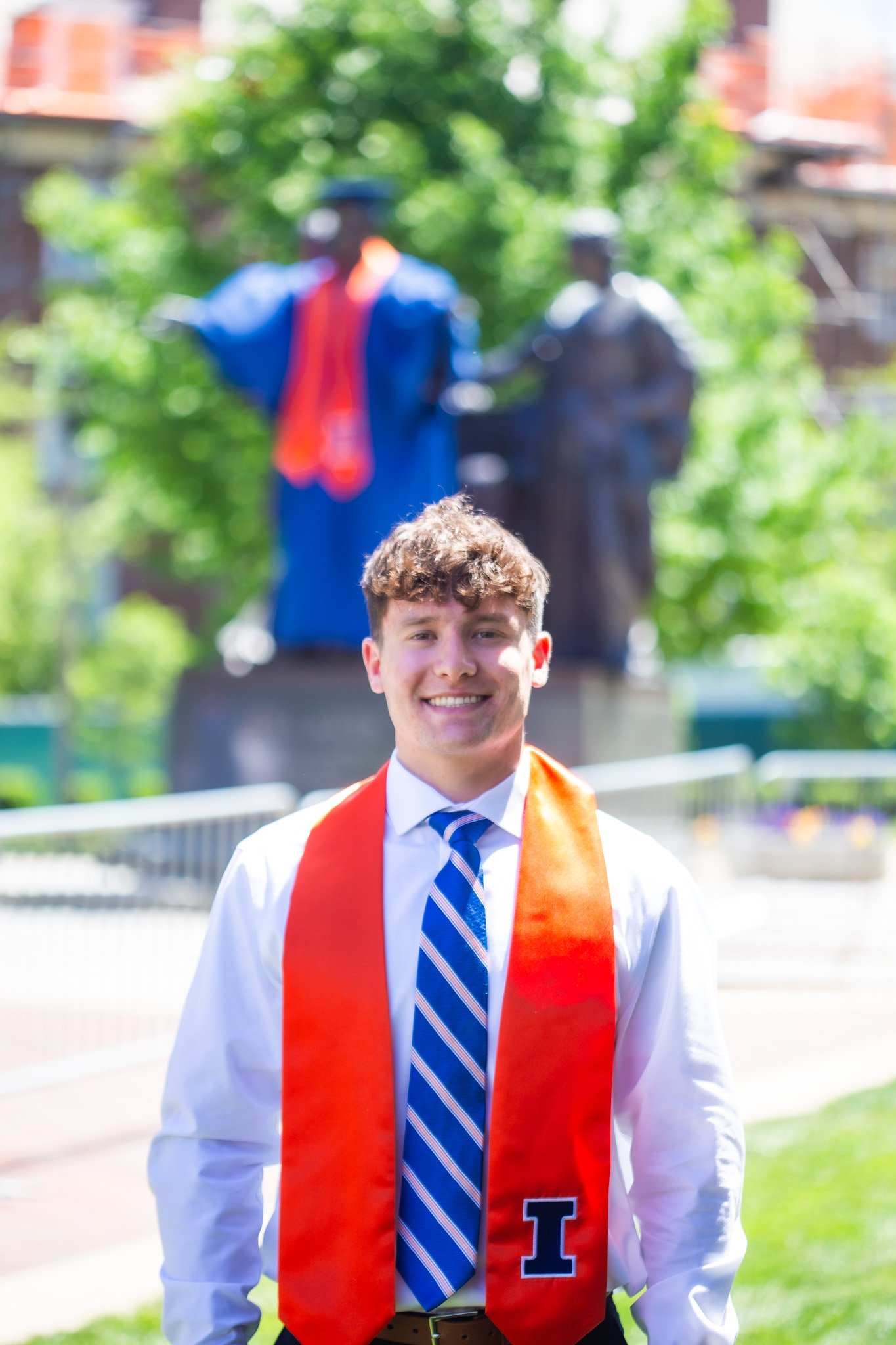 A young man with curly brown hair wearing a white shirt, blue striped tie, and red graduation stole with the letter 'I' on it, smiling outdoors in front of a background with a monument and greenery.