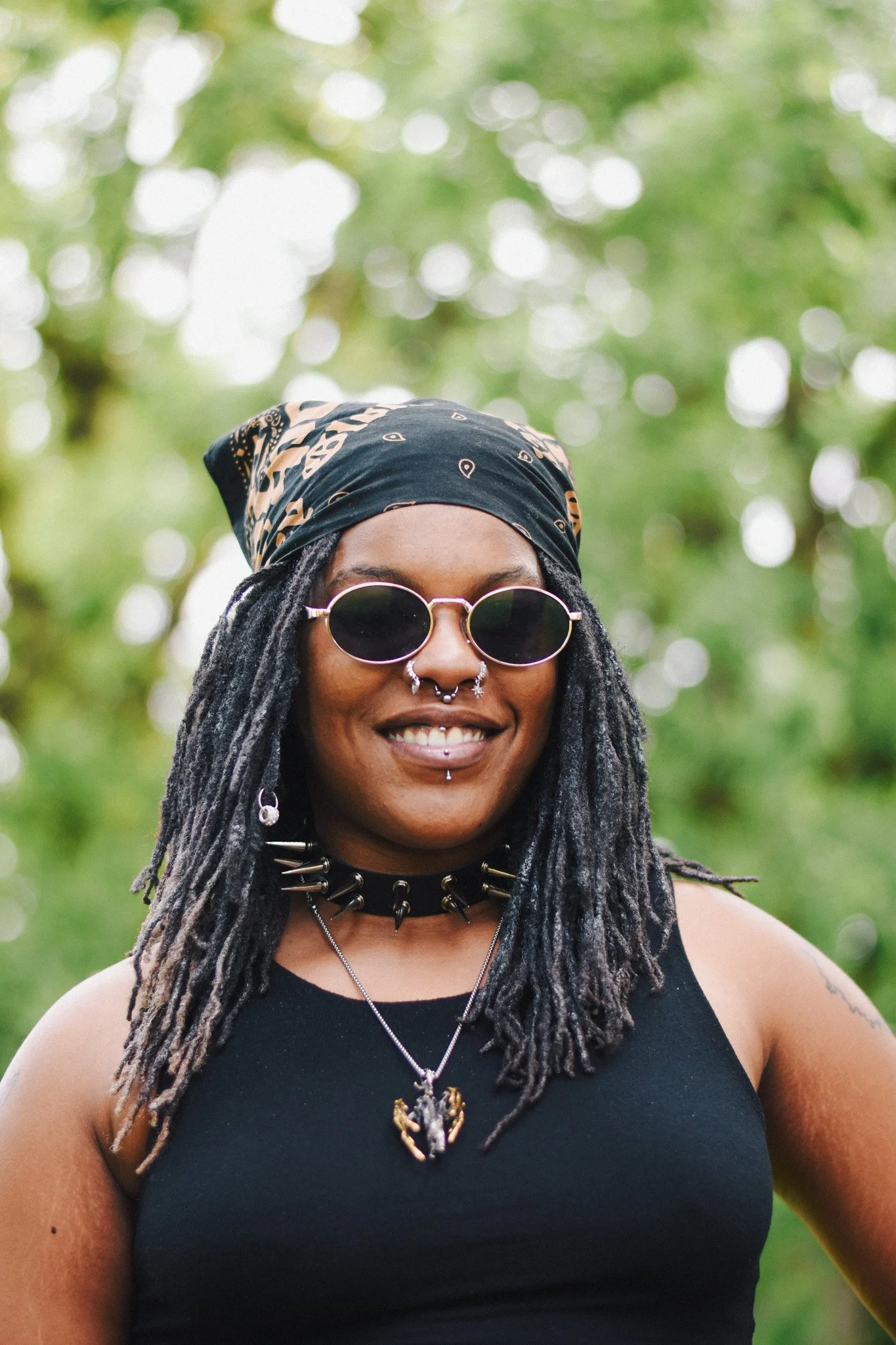 A smiling person with long dreadlocks wearing sunglasses, a black bandana, and various jewelry in an outdoor setting with green trees and bokeh background.
