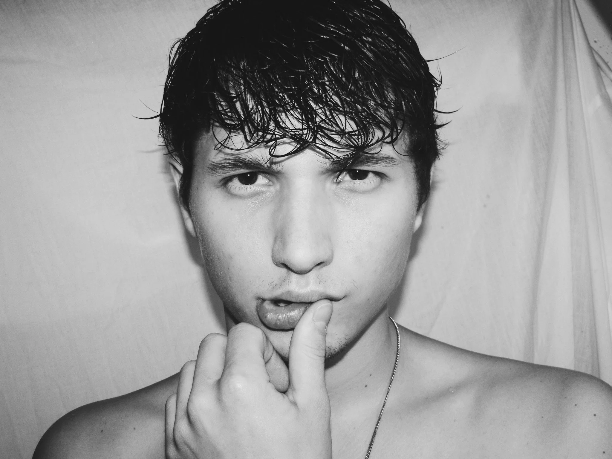 A black and white portrait of a young man with wet, curly hair looking directly at the camera. He is touching his lips with his finger and is shirtless, wearing only a necklace.