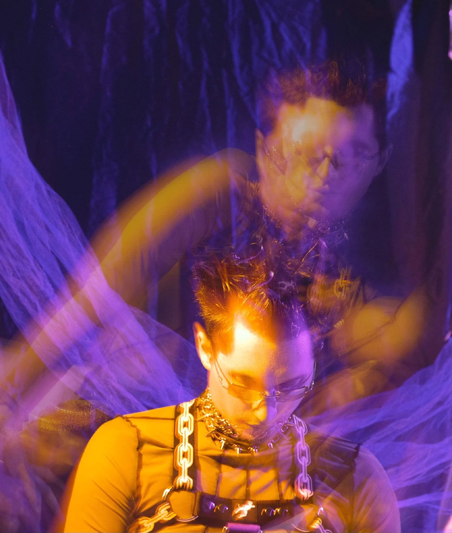 A person wearing a leather jacket, chains, and accessories illuminated by purple and yellow lights, with abstract background effects.