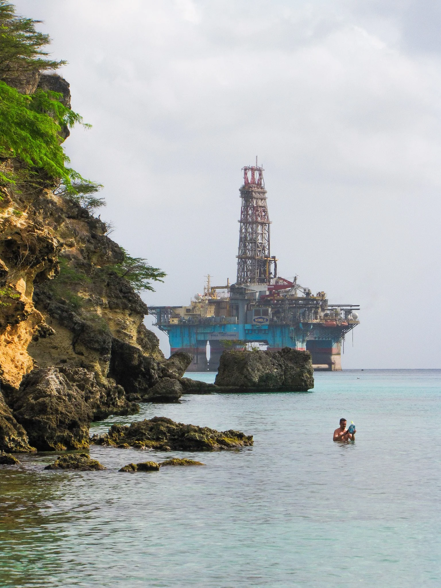 A man holding a water bottle while standing in the water near rocky shoreline with an offshore oil rig in the background.