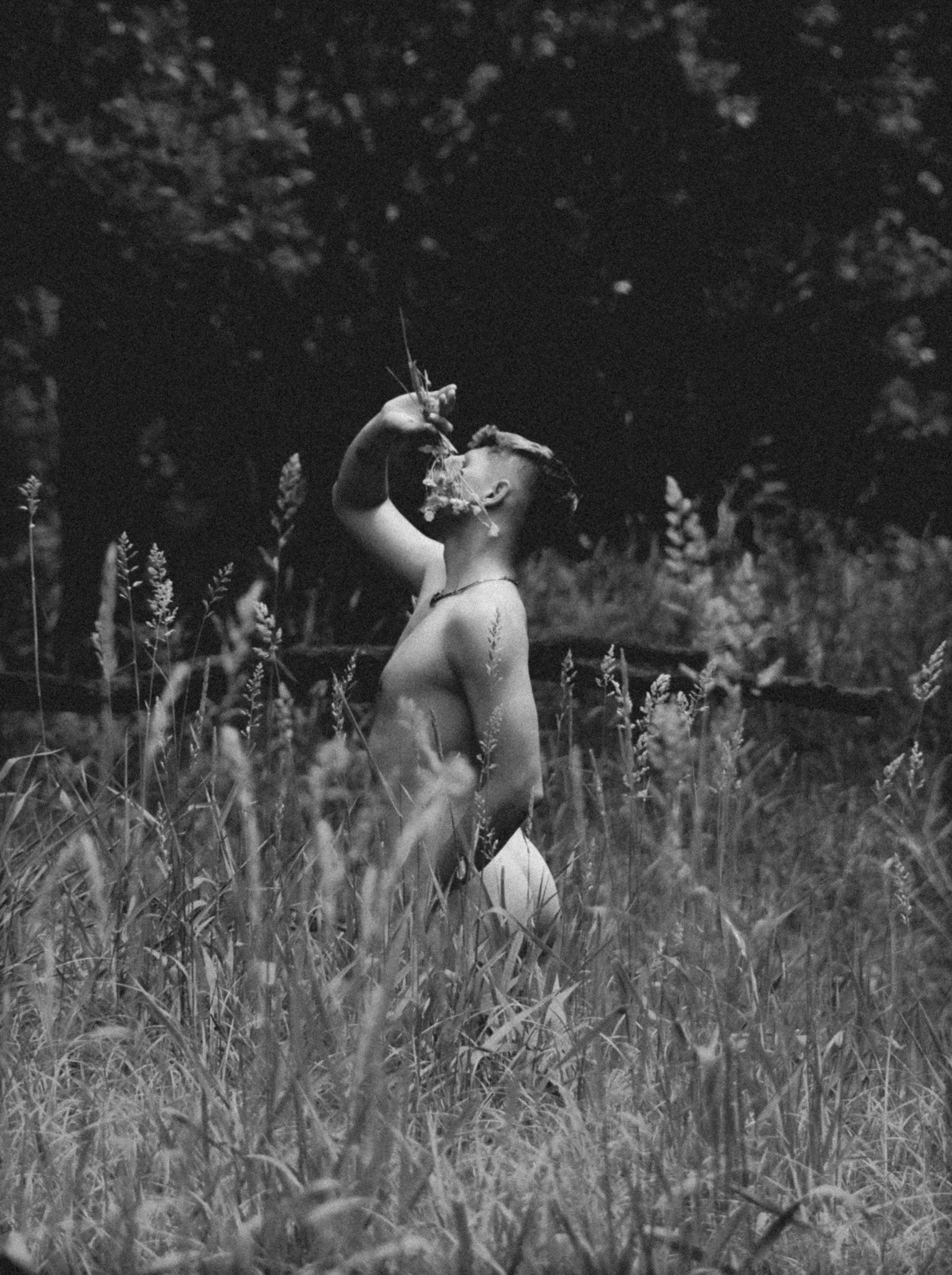 A person kneeling in a field of tall grass and wildflowers, holding a bunch of flowers to their face, in black and white.