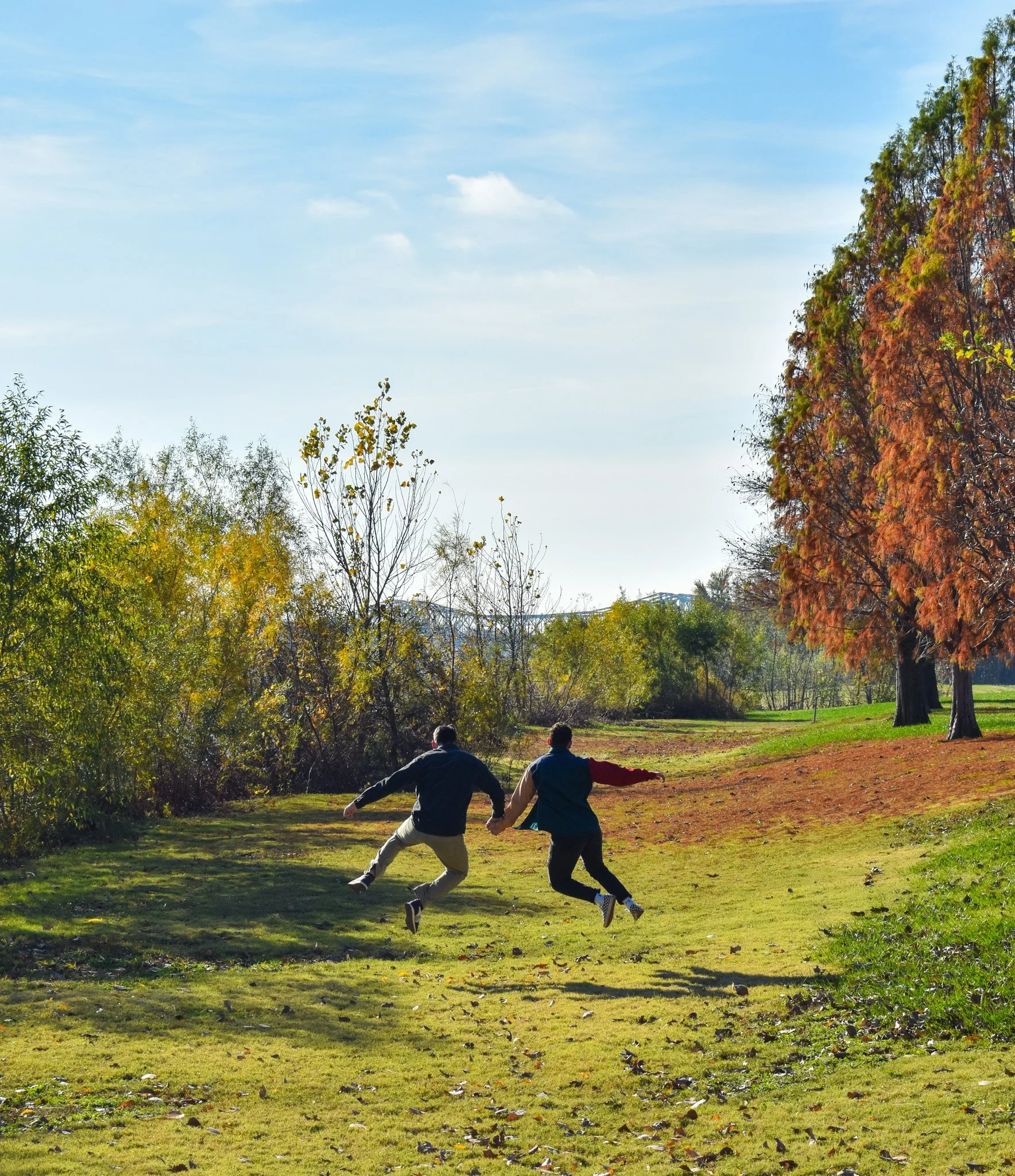 Two people are running across a grassy park with trees showing fall colors. The sky is clear and blue.