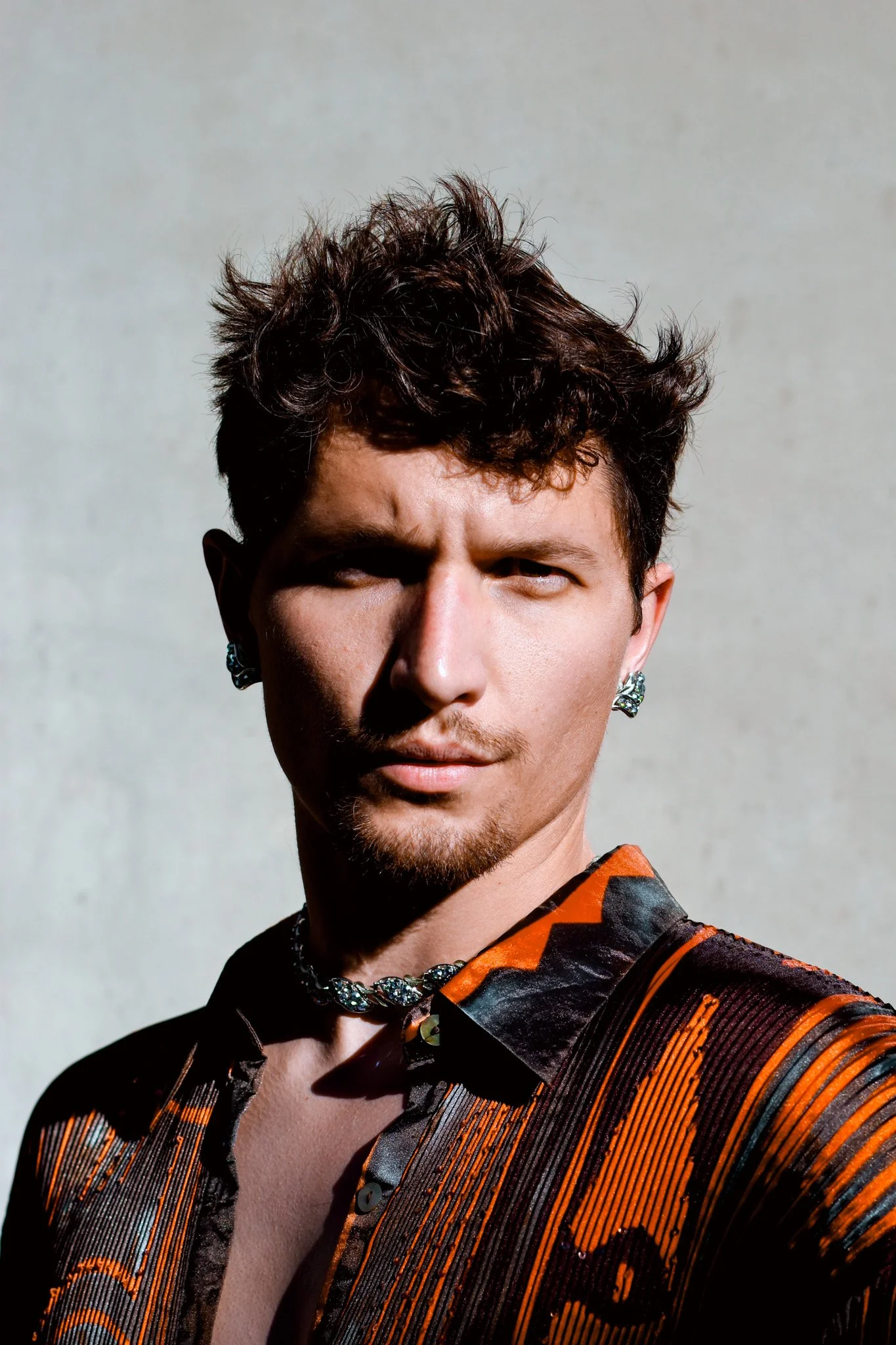 Close-up portrait of a person with short, curly dark hair, wearing earrings, a necklace, and a vibrant patterned shirt with orange, black, and gray colors, standing against a plain light background.