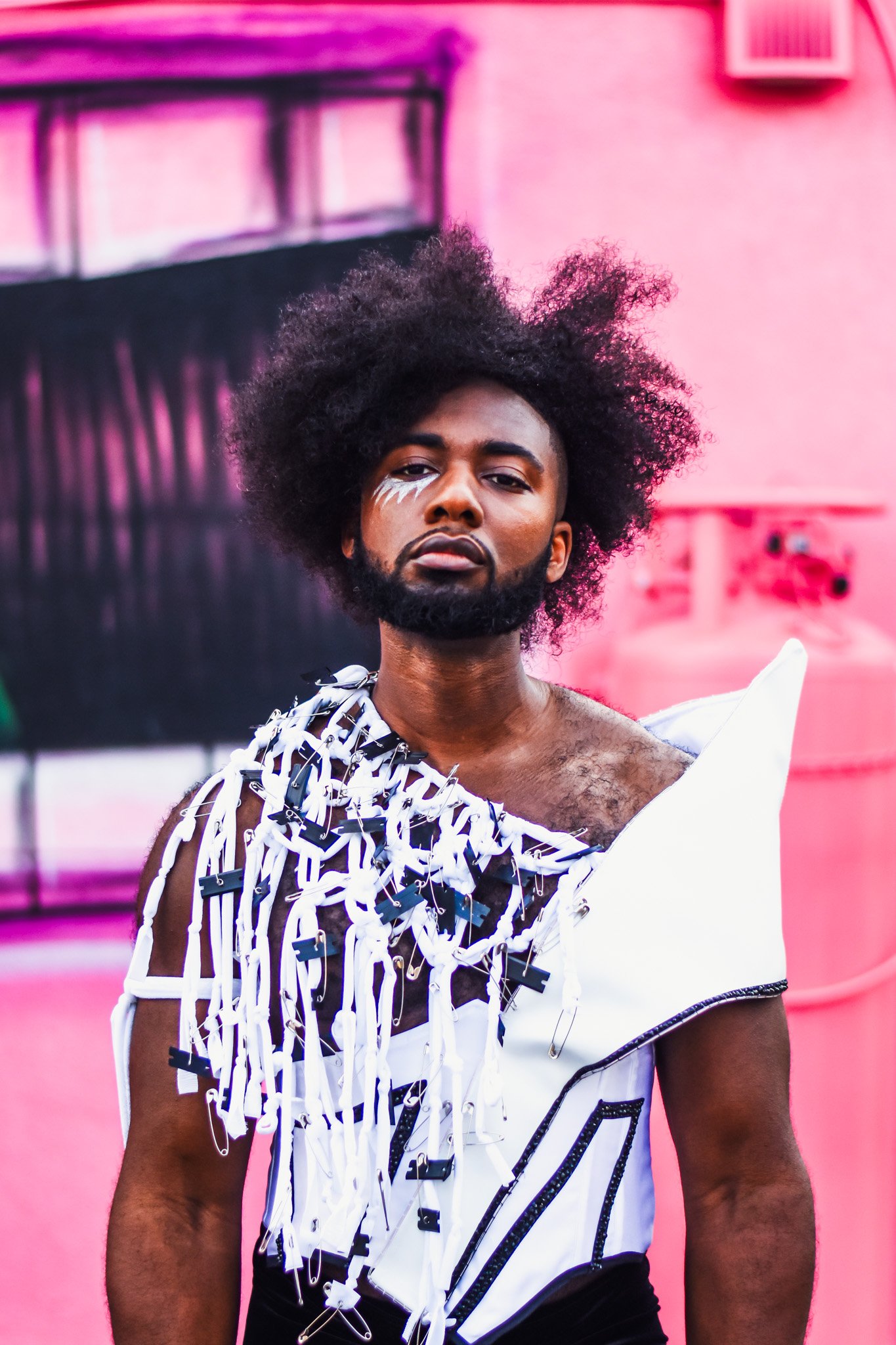 A person with dark skin, a full beard, and a voluminous afro hairstyle standing in front of a pink background, wearing an avant-garde white and black outfit with numerous plastic clips and straps.