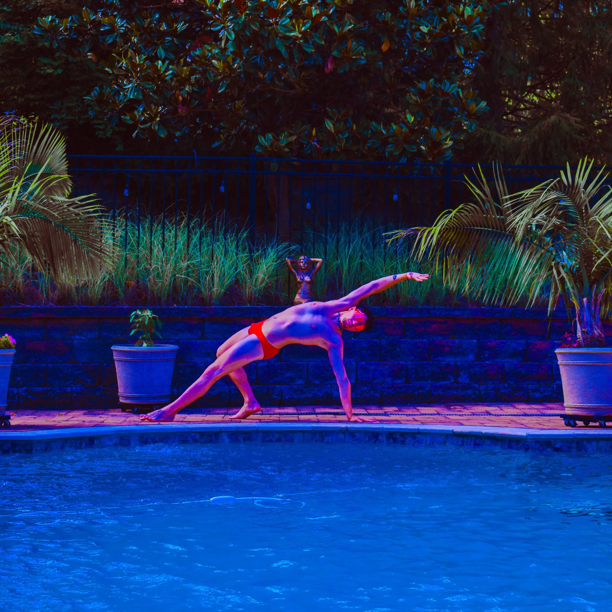 A woman performs a yoga pose near a pool with palm plants, a fence, and trees illuminated by colorful lights at night.