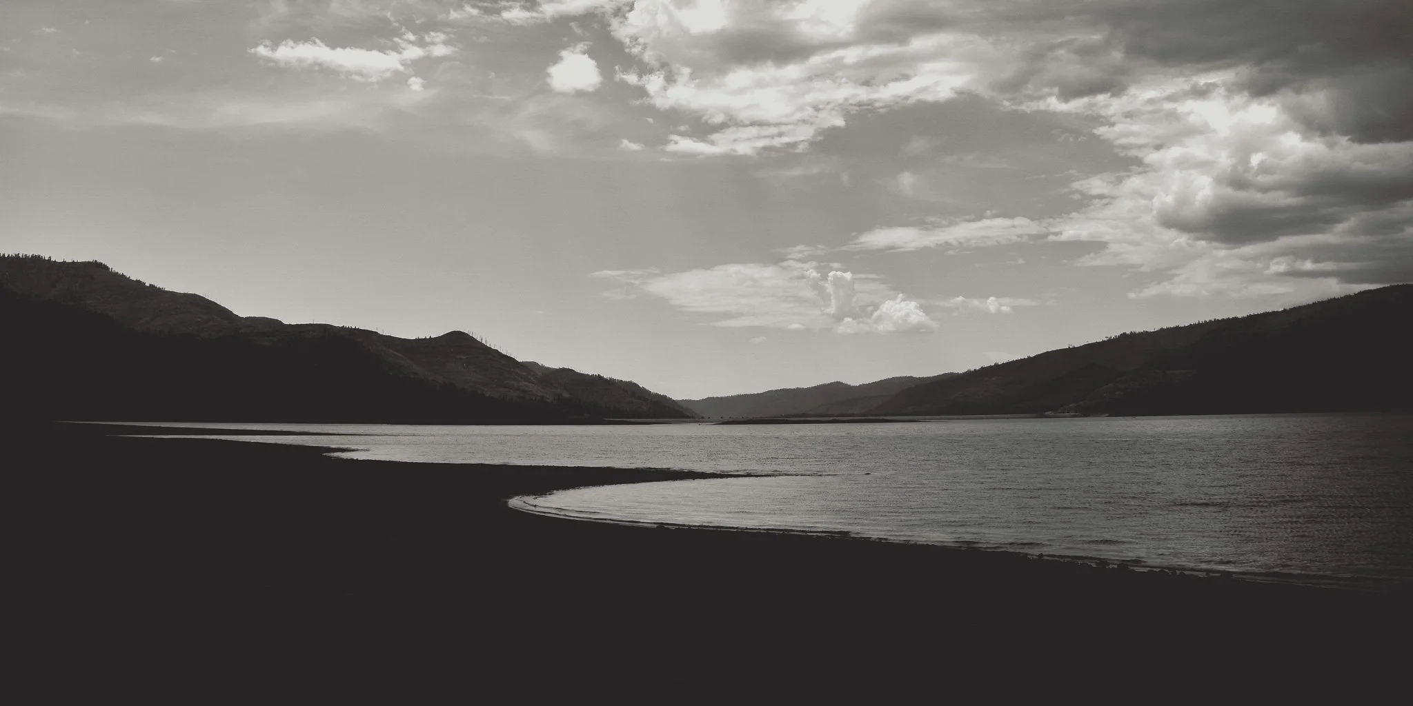 A black-and-white landscape photo of a river or lake with mountains in the background and cloudy sky.