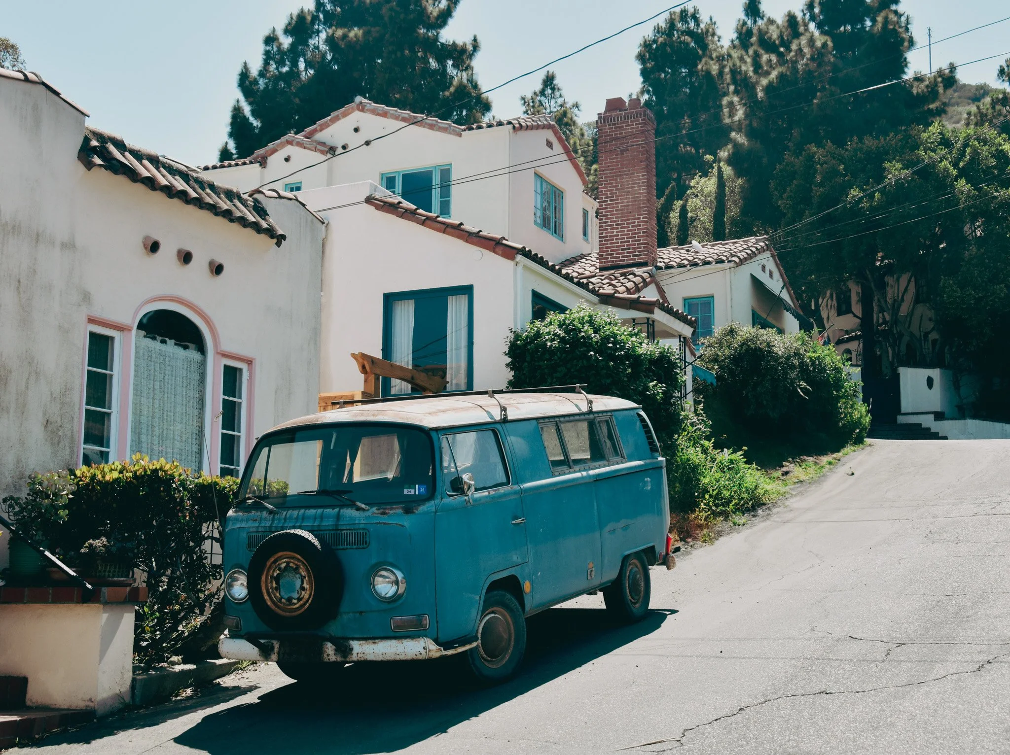 An old blue Volkswagen van parked on a street in front of a white house with blue windows and red roof tiles. There is a bush and flower pot in the foreground, and trees and power lines in the background.