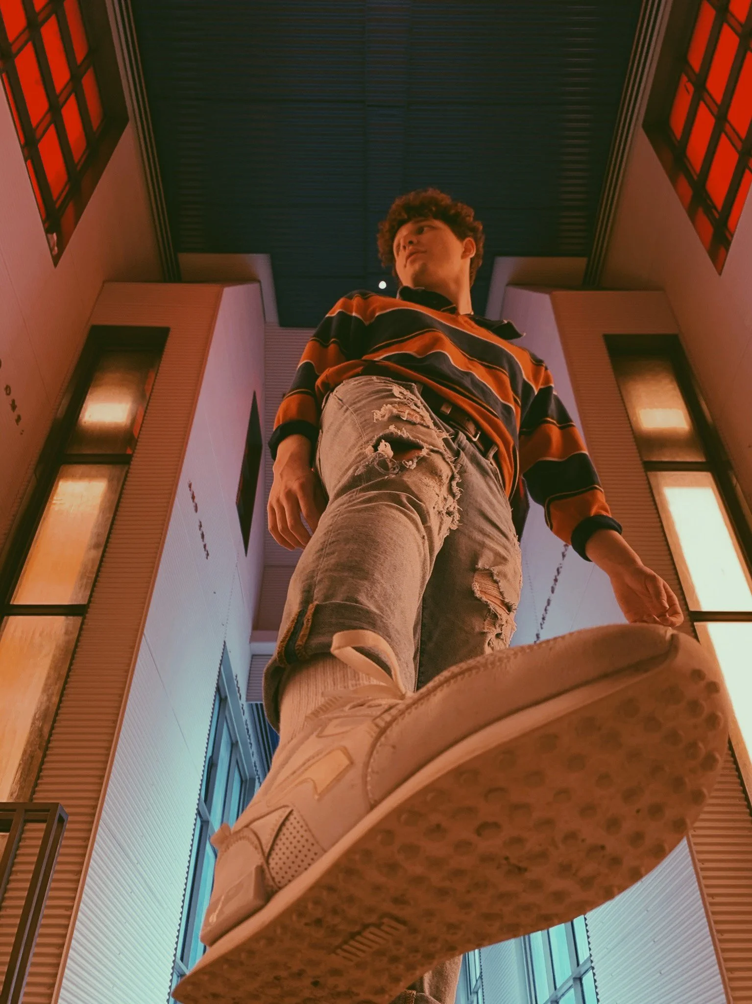 Low-angle view of a young man standing indoors, wearing a striped shirt and distressed shorts, with a modern interior background featuring wooden panels and large windows.