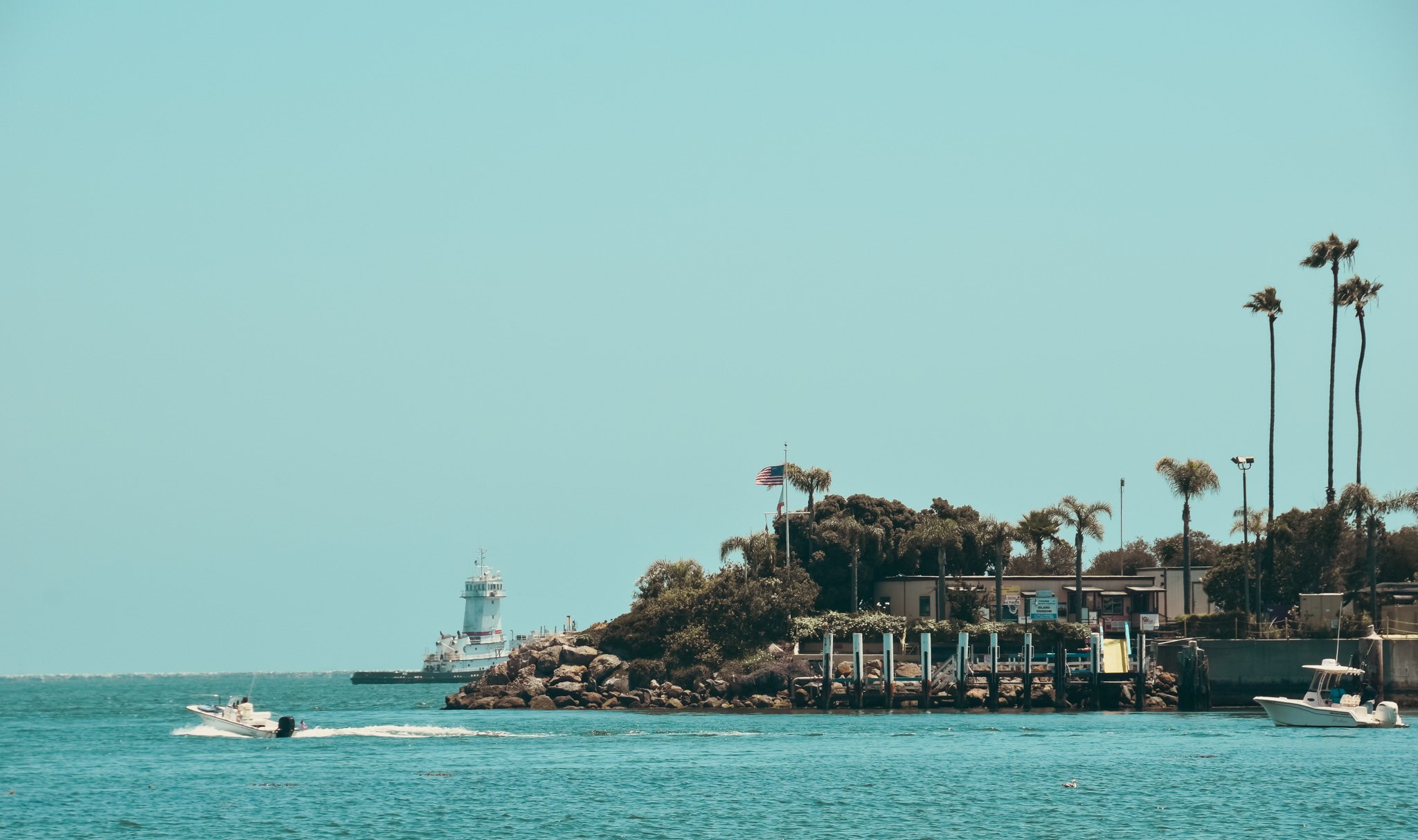 A coastal scene with a lighthouse, boats in the water, and palm trees against a clear sky.