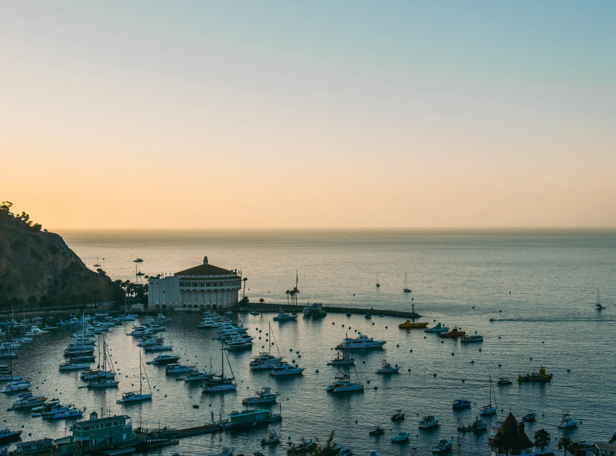 A harbor filled with numerous white boats and yachts, with a large white building, possibly a hotel or clubhouse, near the waterfront. The background features the ocean and a sky transitioning from light to darker colors at sunset.