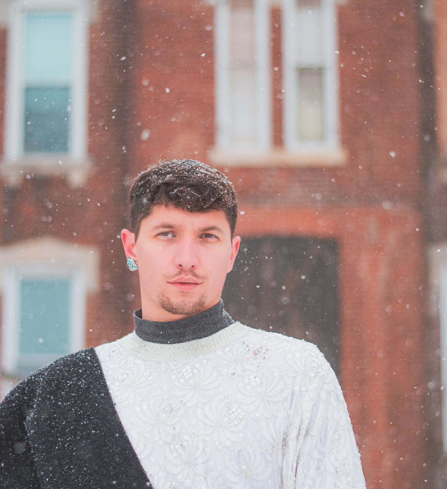 Close-up of a young man with short dark hair, wearing a white lace top and black outerwear, standing outside during snowfall. The background shows a brick building with windows.