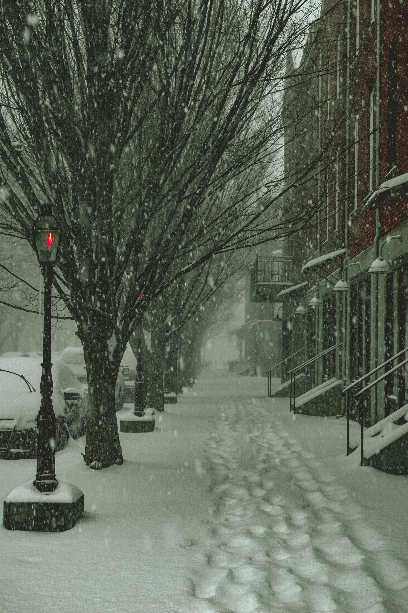 Snow-covered sidewalk in a neighborhood with trees and residential buildings, footsteps in the snow, and a streetlamp with a red traffic signal.