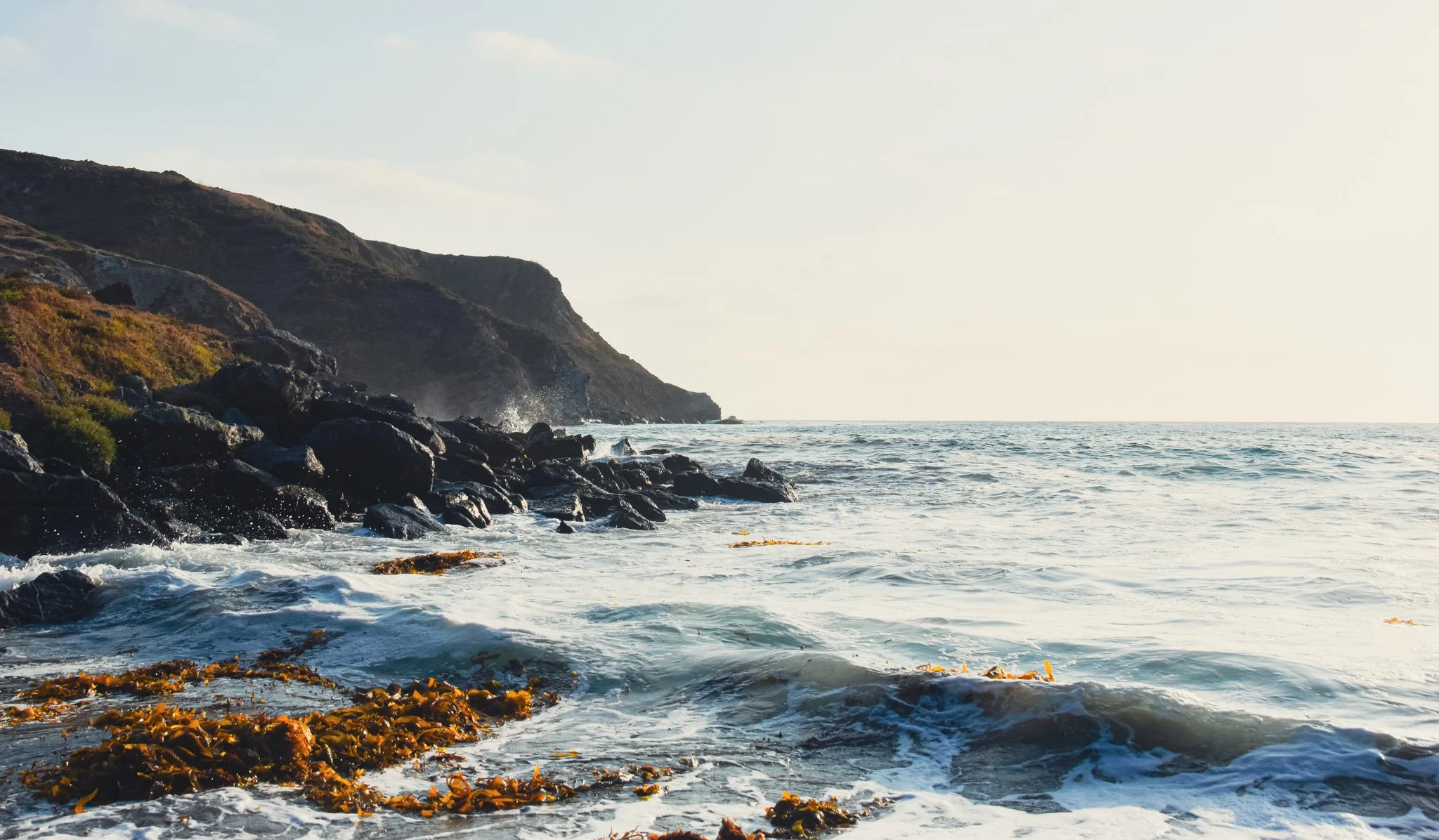 A rocky shoreline along the ocean with waves crashing onto the rocks, a hillside covered with vegetation, and distant cliffs under a bright sky.