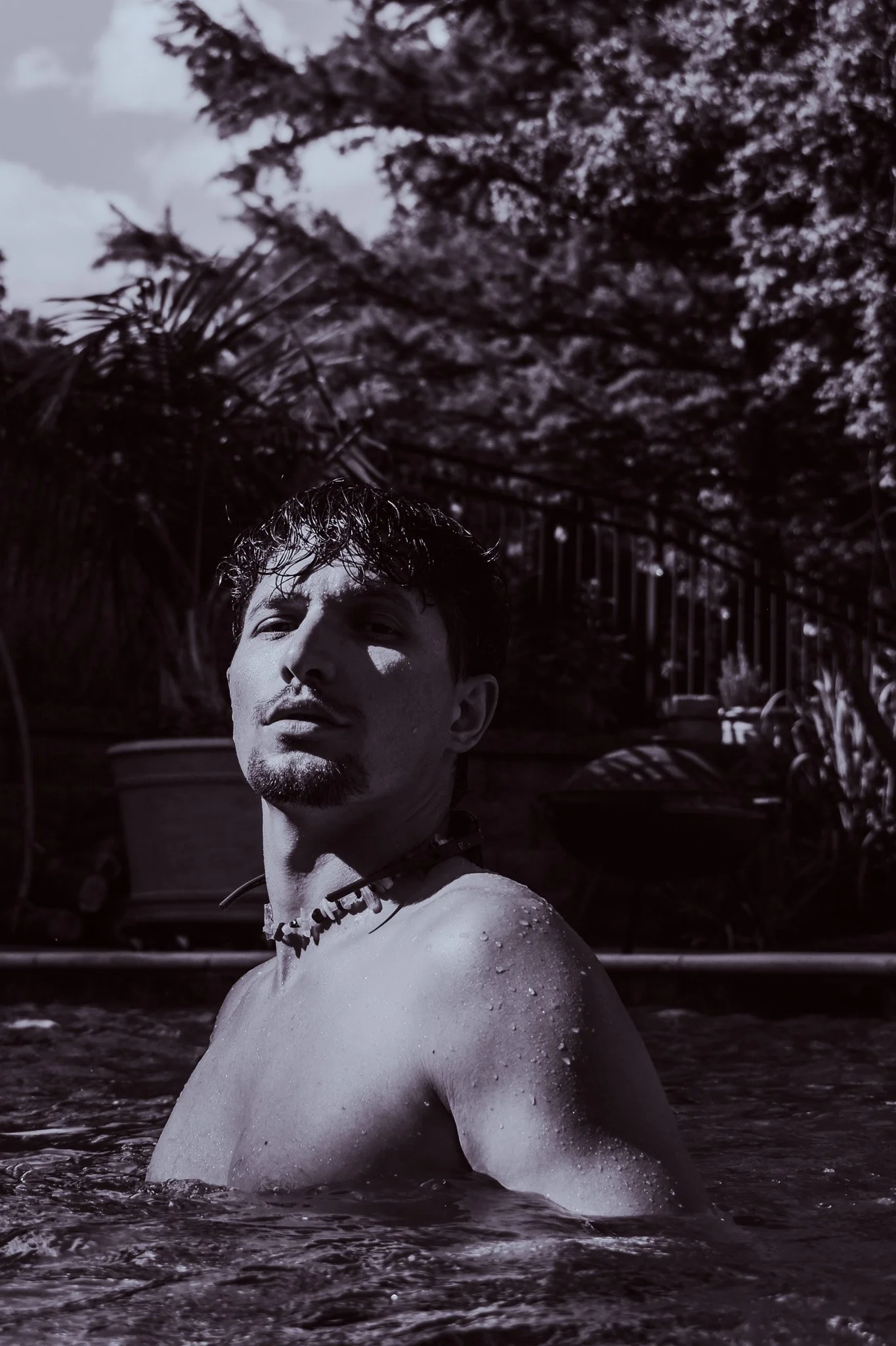 Black and white photo of a person with wet hair neck and shoulders in a swimming pool, with water droplets on skin, surrounded by trees and foliage.