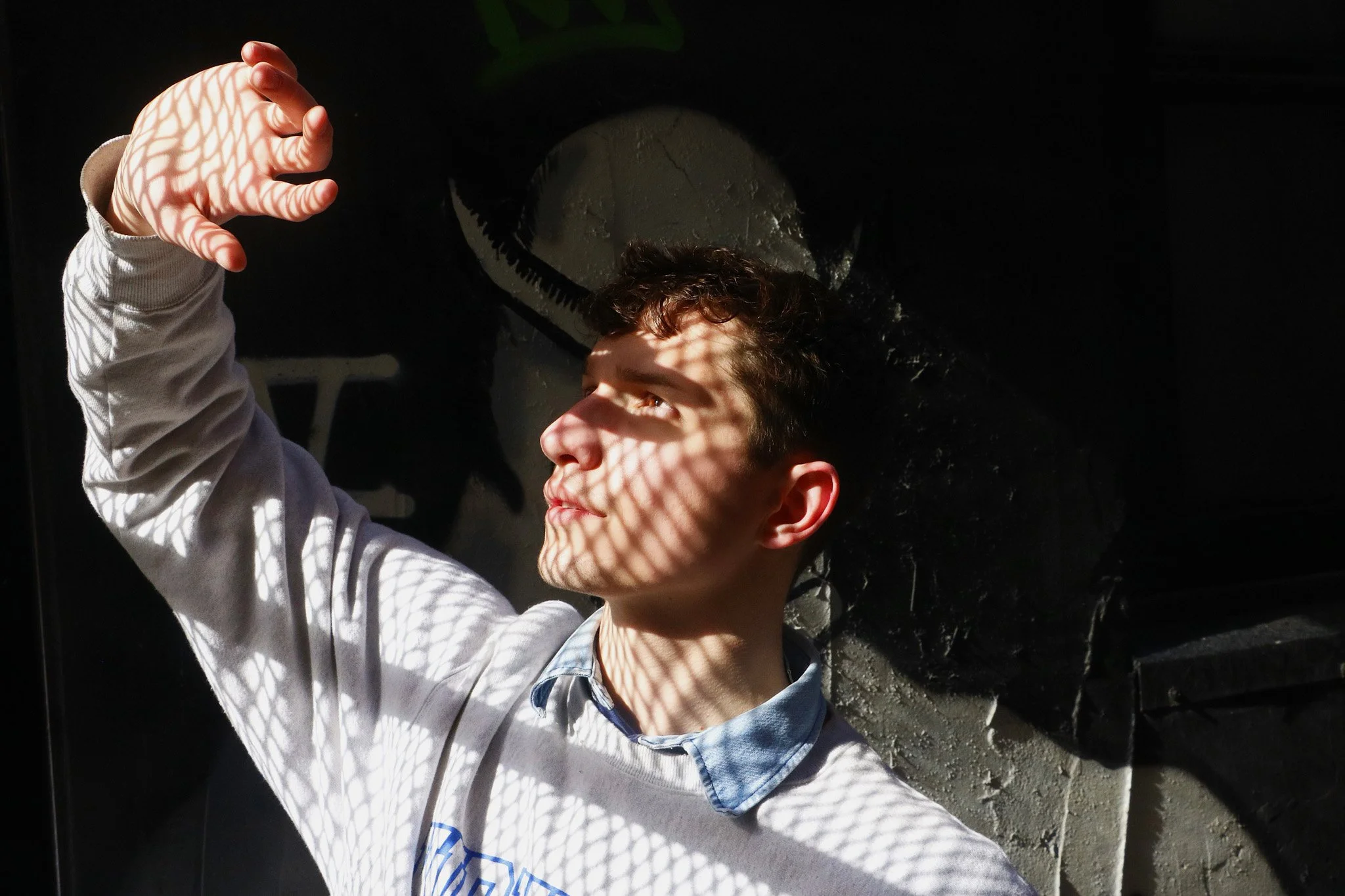 A young boy with curly brown hair and light skin looking up, with sunlight casting patterned shadows through a wire or lattice, creating a geometric shadow on his face and hand. He is wearing a light-colored hoodie.