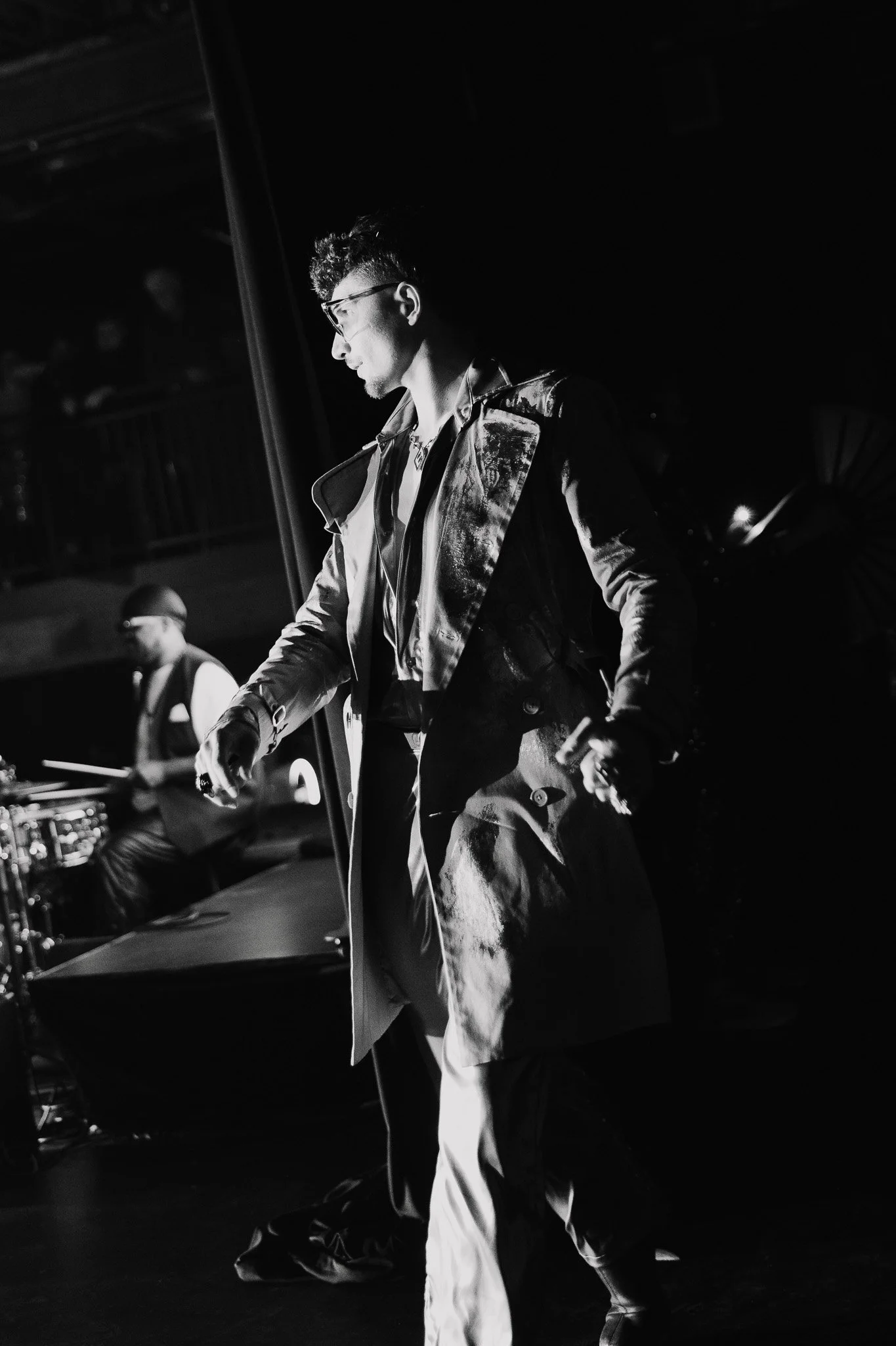 A black and white photo of a man with glasses and curly hair, wearing a shiny leather jacket and light-colored pants, walking or moving in a dark environment with another person in the background.