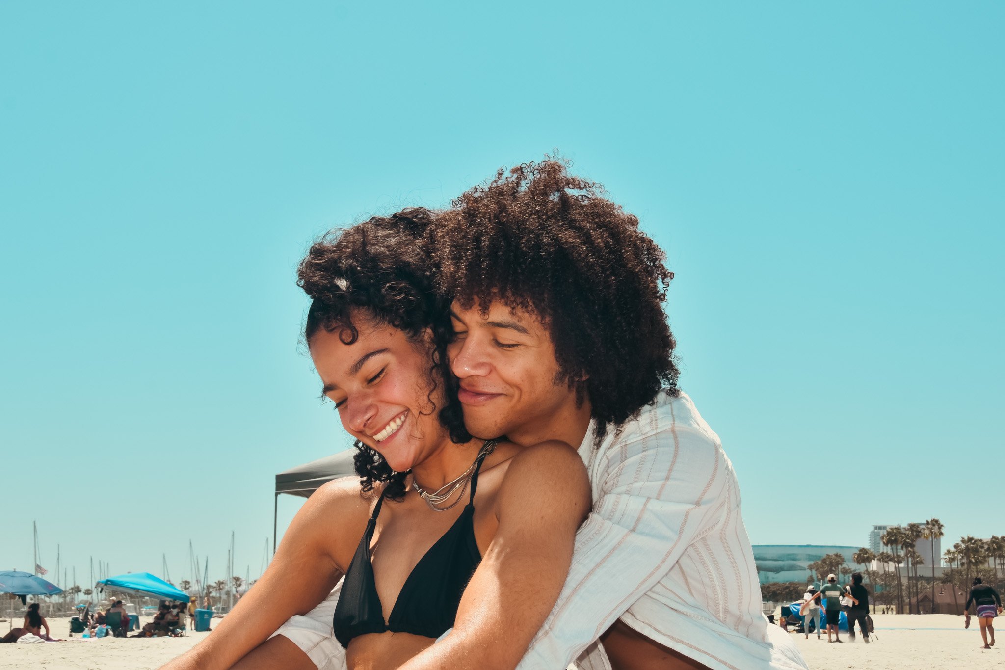 Two people, a man and a woman, hugging and smiling at the beach under a clear blue sky.