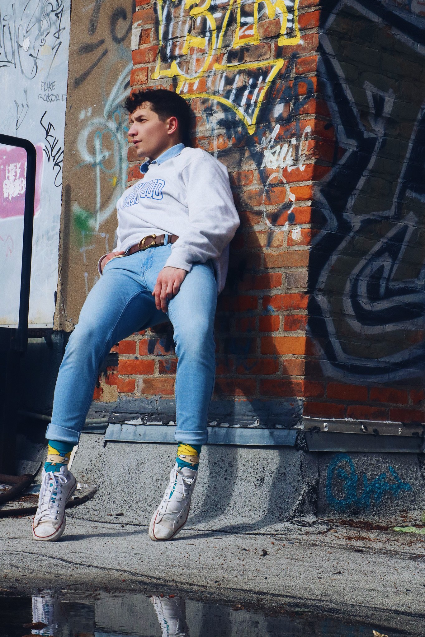 A young man with curly hair leaning against a brick wall covered in colorful graffiti in an urban alley. He is wearing a white sweatshirt, light blue jeans, and colorful sneakers with yellow socks. He looks to the side with a serious expression, and 